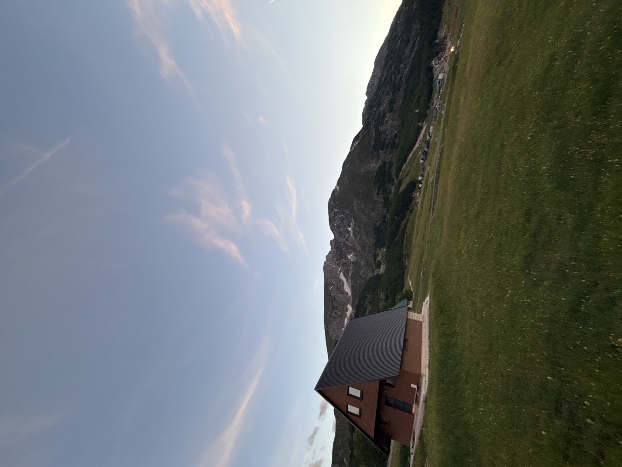 In the city of Virak, Montenegro, a pitched-roof brown wooden cabin with two square windows and a dark gray metal roof sits on a grassy slope. The cabin is angled towards the left side of the frame. Behind the cabin, a mountainous landscape rises with dark green trees covering the lower slopes, transitioning to rocky, snow-dusted peaks under a pale blue sky with wisps of pink clouds. In the distance, several small buildings and structures are visible scattered along the mountainside. The foreground is dominated by a field of green grass, dotted with small yellow flowers. The lighting suggests it is either dawn or dusk.