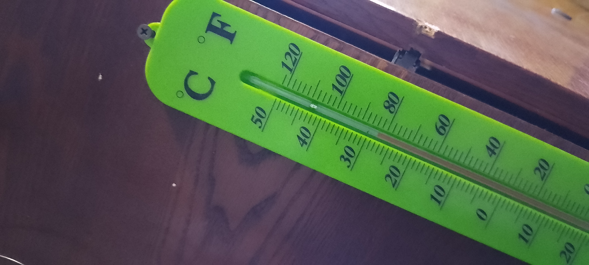 The image shows a close-up, angled view of a bright green thermometer with both Celsius and Fahrenheit scales. The thermometer is mounted on a dark wooden surface. The Celsius scale is on the left and shows markings for 0, 10, 20, 30, 40, and 50 degrees. The Fahrenheit scale is on the right and shows markings for 20, 40, 60, 80, 100, and 120 degrees. A red liquid column is visible within the thermometer's tube, indicating a temperature reading between 40 and 50 degrees Celsius, which corresponds to approximately 100 to 120 degrees Fahrenheit. The top of the thermometer has a small screw securing it to the wooden surface.