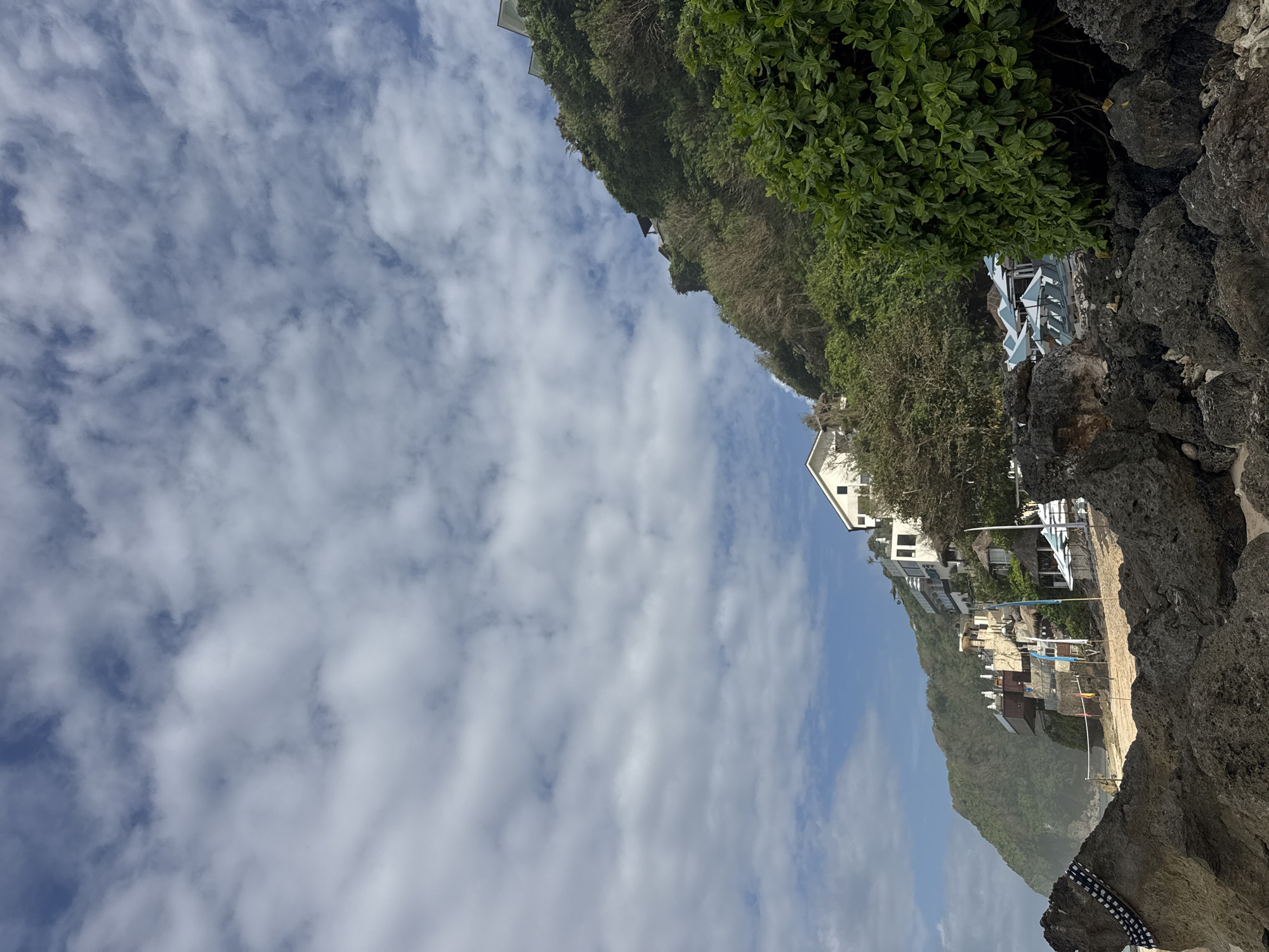 The image shows a view in Kuta Selatan, Indonesia. The sky is partially cloudy with a mix of blue and white clouds. The coastline features cliffs covered with lush green vegetation and buildings perched on the cliffs. A series of structures and buildings are visible down the cliffside. Rocks and boulders are in the foreground.