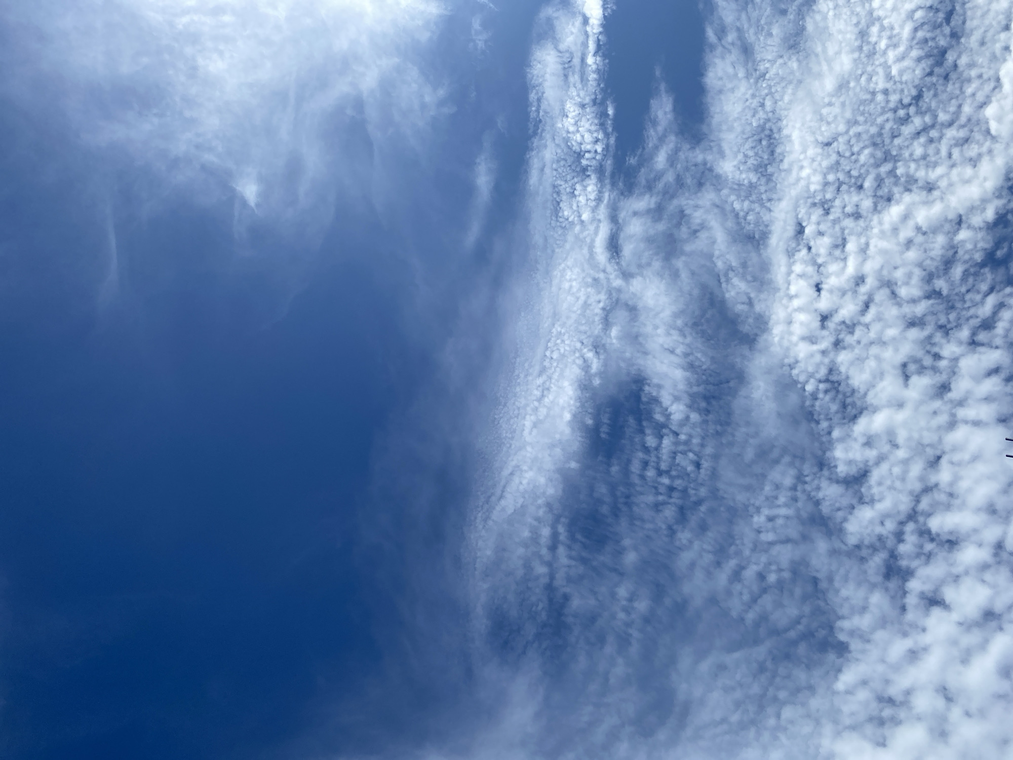 The image is a wide shot of a bright blue sky filled with white, wispy clouds. The clouds are scattered across the sky, with some appearing thin and elongated, while others are more clumped together, forming a textured pattern. The sun is not directly visible, but its presence is indicated by a bright white glow in the upper right portion of the frame, which illuminates the clouds from behind and creates a lens flare effect. The vast expanse of the sky suggests an outdoor scene, likely during the day, with no visible signs of human activity or identifiable landmarks.