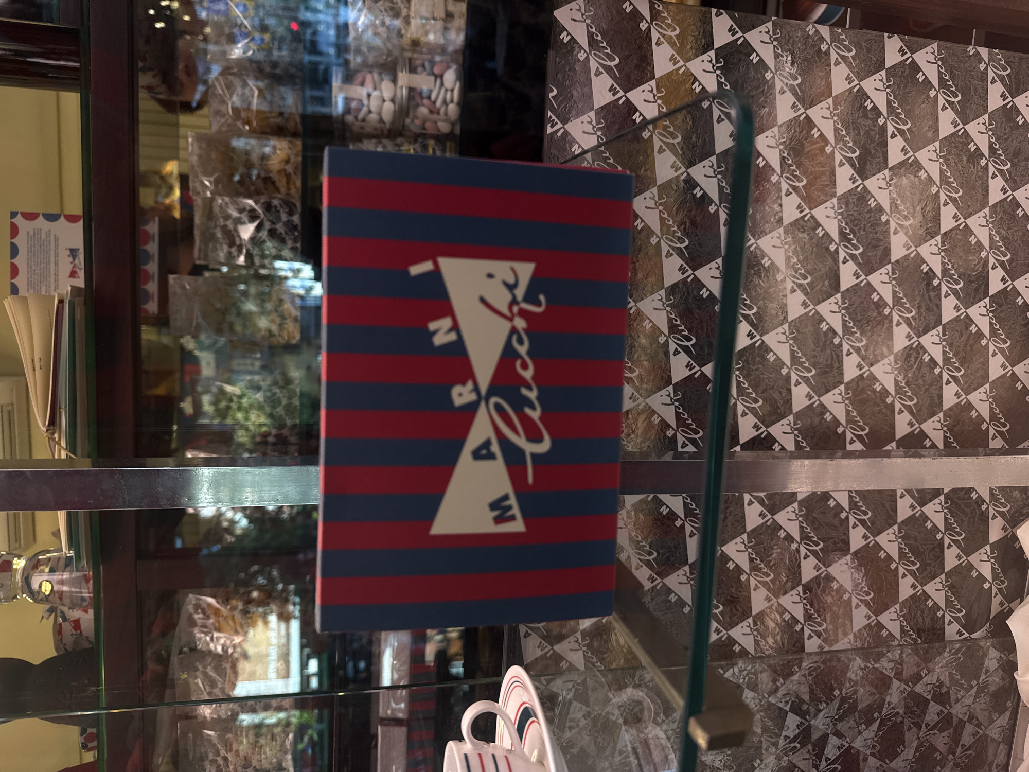 The image captures a shop display in Milan, Italy, showcasing merchandise. The dominant element is a box with horizontal red and blue stripes, emblazoned with the name "MARNI Lucchi" in cursive. Adjacent to it is another box patterned with a repeating "Lucchi" design. A shelf with various packaged goods is visible through the glass display. In the foreground, parts of some white tableware, possibly cups and saucers, are discernible. The scene is contained within a glass cabinet.