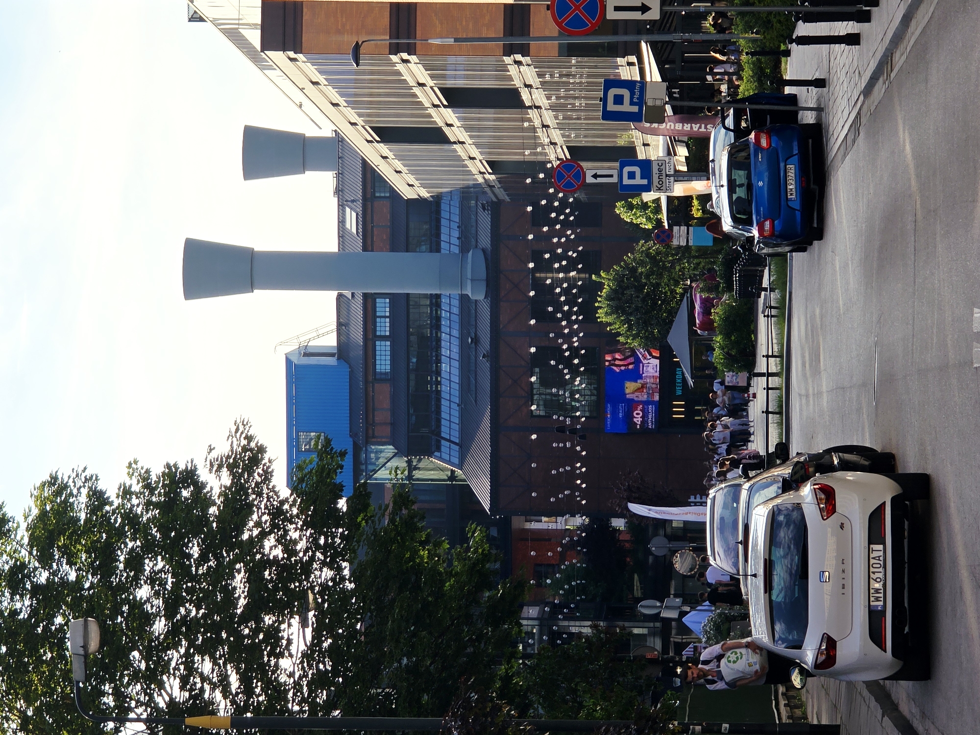 The image captures a street scene in Warsaw, Poland, likely in the area of Plac Grzybowski, known for its revitalized industrial architecture. The foreground features two cars parked on the right side of the road: a white SEAT Ibiza with license plate WW 610AT and a blue Suzuki Swift with license plate WM 9377R. The Suzuki has its driver's side door open. A man carrying a large white bag with recycling symbols walks along the sidewalk on the left.
Behind the cars, several people are visible walking on a sidewalk or promenade. The background showcases a striking blend of modern and industrial architecture. Prominently featured are two large, grey cylindrical structures resembling industrial chimneys, part of a repurposed factory building. This building exhibits exposed brickwork and large glass windows. Adjacent to it is a modern glass-facade building. String lights are suspended across the street between the buildings, suggesting a festive or inviting atmosphere. Several traffic signs are visible, including parking signs and a "Koniec Stref Ruchu" sign, indicating the end of a traffic zone. The sky is bright and appears to be late afternoon or early evening.