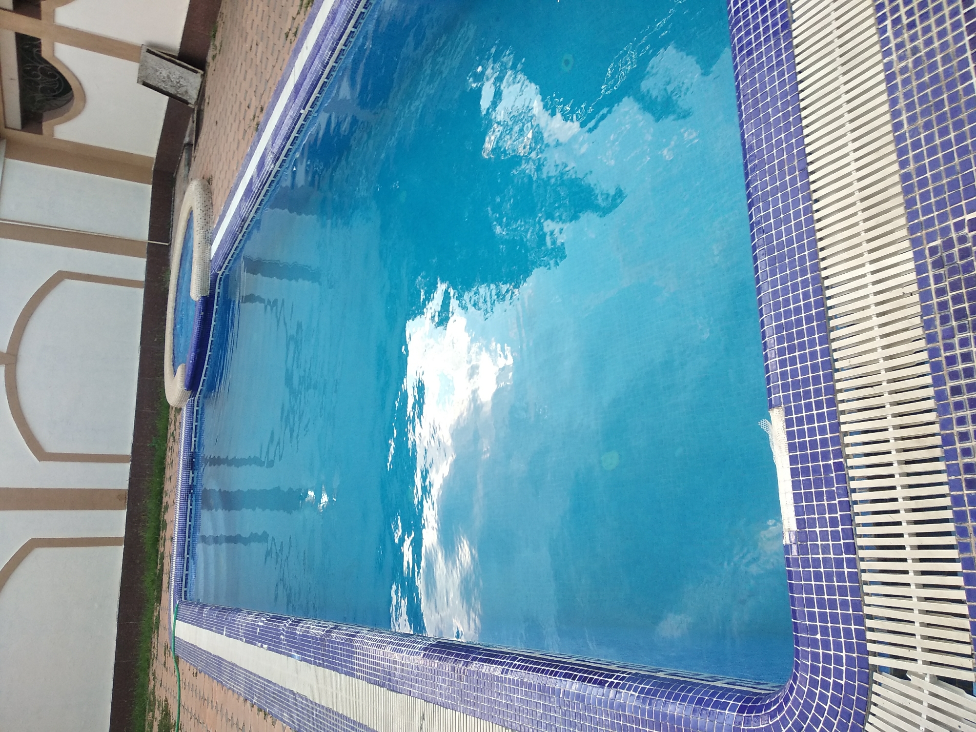 An empty swimming pool is shown in the city of Charvak, Uzbekistan. The pool is rectangular and filled with clear blue water that reflects the sky and clouds. The edge of the pool is lined with blue and white mosaic tiles, and a white grate runs along the perimeter for drainage. To the left of the pool, there is a paved area with brick-like stones and some scattered grass. In the background, a light-colored wall with arched decorative elements can be seen. To the right, a small circular jacuzzi with a stone border is partially visible, and a wooden board leans against the wall. The overall scene suggests an outdoor recreational area.