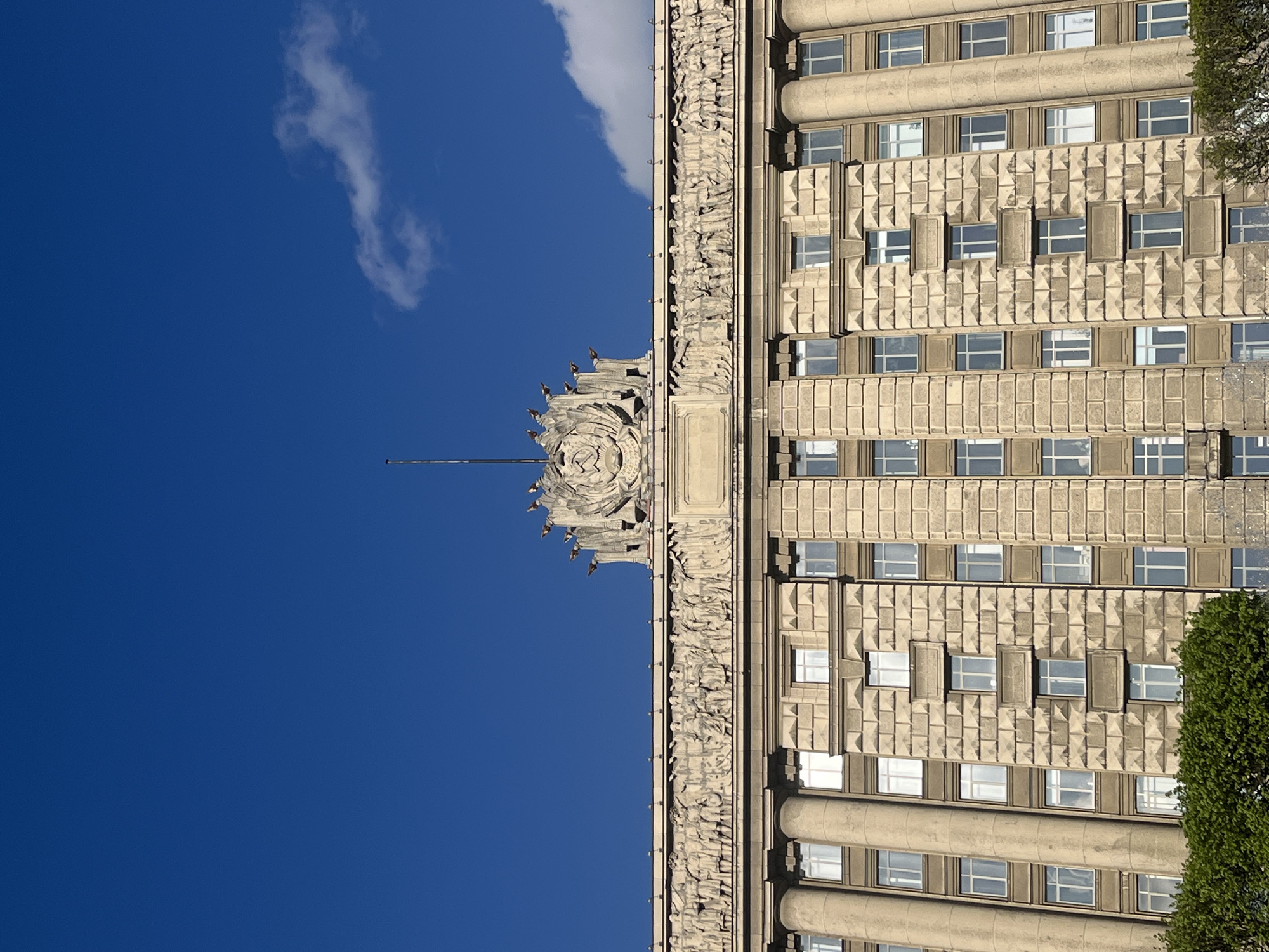 The image shows a portion of the Saint Petersburg Legislative Assembly building in Russia, viewed from below. The building features a classical facade with repeating windows, decorative stone elements, and a central crest at the top. The view includes a deep blue sky with a wispy cloud. The structure is rendered in a tan color, contrasting with the clear sky. A tree with green foliage is also visible on the right.