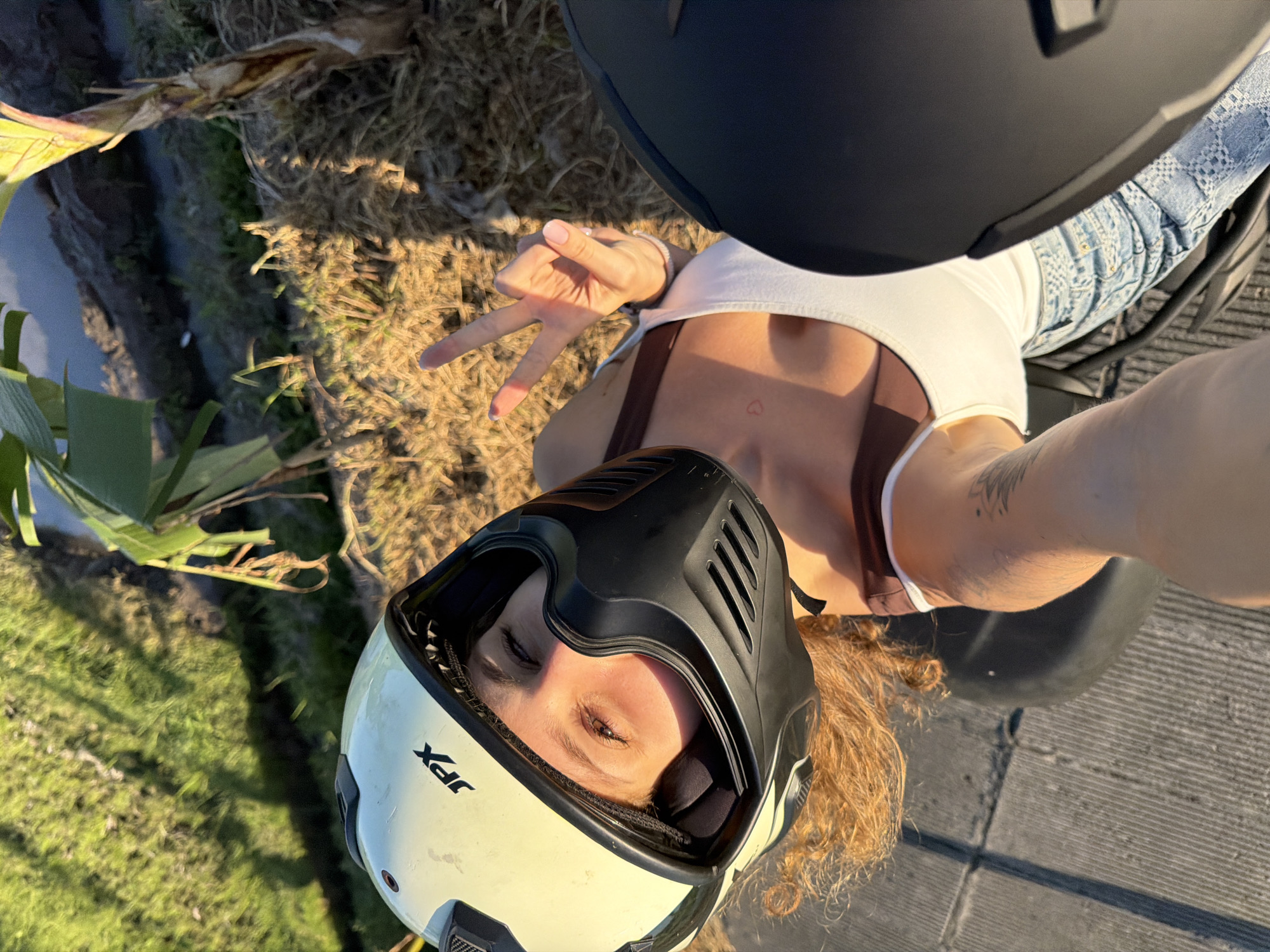 A woman wearing a white and black motorcycle helmet, a white tank top, and denim pants sits on a motorcycle in North Kuta, Indonesia. She is facing upwards, holding her arm up in a selfie pose while making a peace sign with her fingers. The environment includes grass, dirt, and vegetation typical of the region.