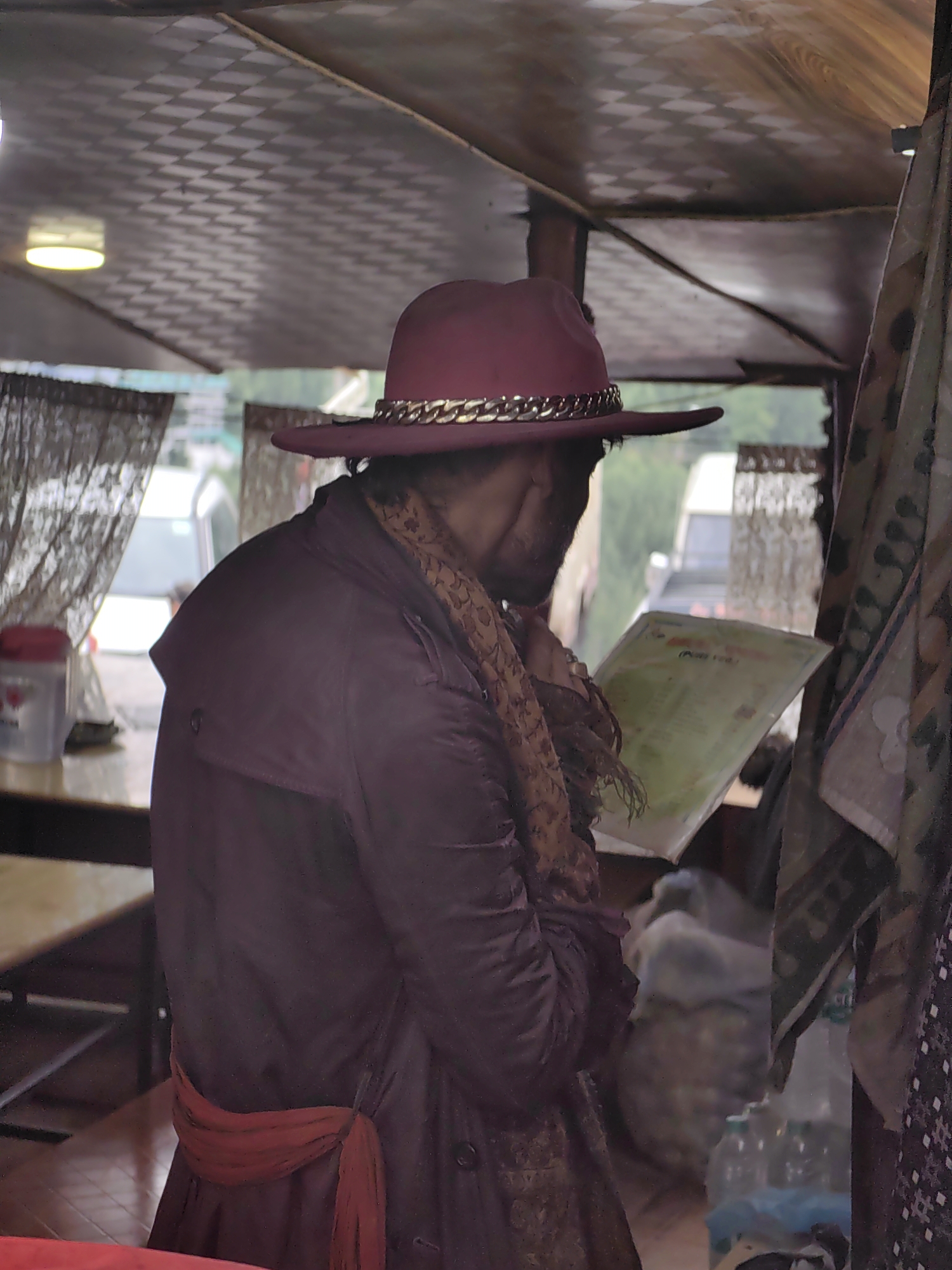 In Vashisht, India, a person wearing a maroon hat adorned with a gold chain and a maroon trench coat is standing inside a covered vehicle. They are holding and reading a green document, possibly a menu or a leaflet. The person is also wearing a patterned scarf around their neck and a sash of reddish fabric around their waist. The interior of the vehicle has a textured ceiling and visible curtains, suggesting it may be a taxi or a small bus. Outside the vehicle, through a window, glimpses of other vehicles and trees can be seen. The overall scene appears to be an indoor setting within a vehicle in Vashisht.