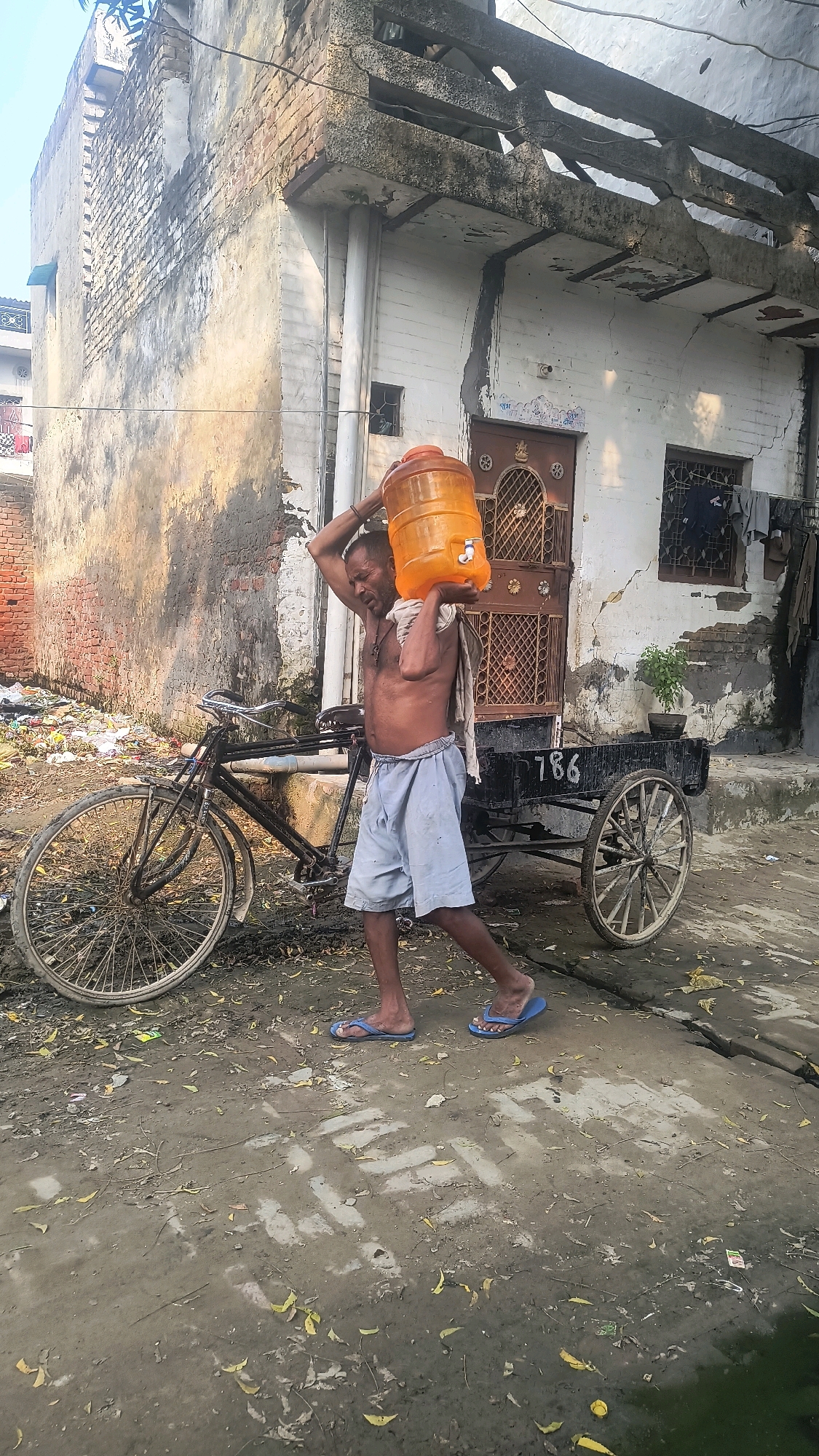 An un-identified man is seen walking and carrying a large orange water jug on his right shoulder. He is wearing light blue shorts and blue flip-flops. He is shirtless and has a cloth draped over his left shoulder. He appears to be in a street in Loni, India, with a bicycle on his left and a cart behind him. The street is unpaved and littered with debris. The buildings in the background are old and in disrepair, with peeling paint and cracked walls. The sky is visible in the upper left corner of the image.