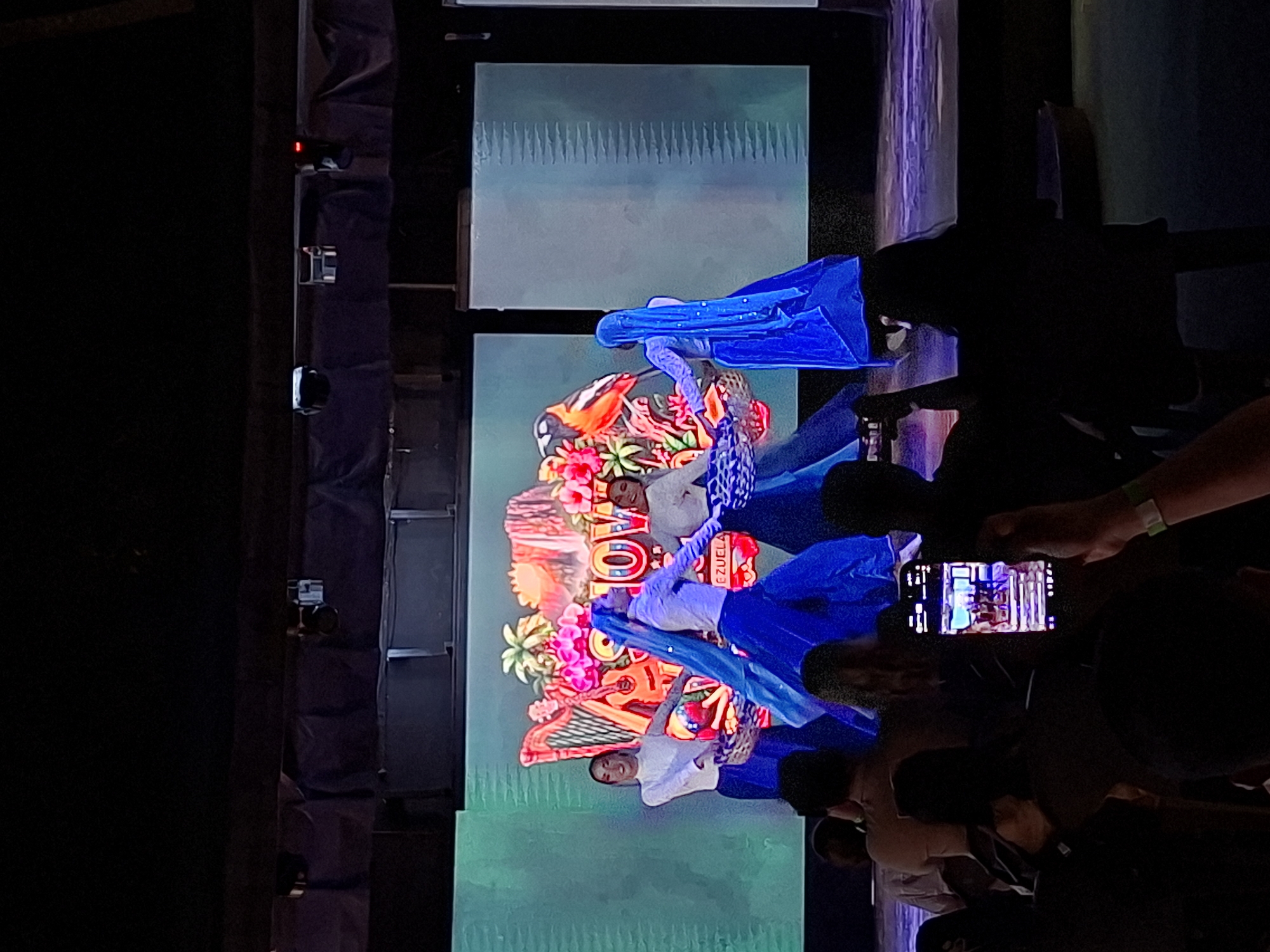This is a photograph taken from a darkened audience looking towards a stage in Porlamar, Venezuela. Four dancers in blue skirts and white tops are performing, holding baskets and some have flowing blue fabric. The backdrop is a large LED screen displaying a vibrant scene with tropical elements, including a harp, flowers, a bird, and what appears to be a waterfall. The word "SHOW" is prominently displayed on the screen, along with "VENEZUELA". The audience members in the foreground are silhouetted, with one person holding up a smartphone to record the performance. The overall lighting suggests an indoor event, likely a cultural performance or show.