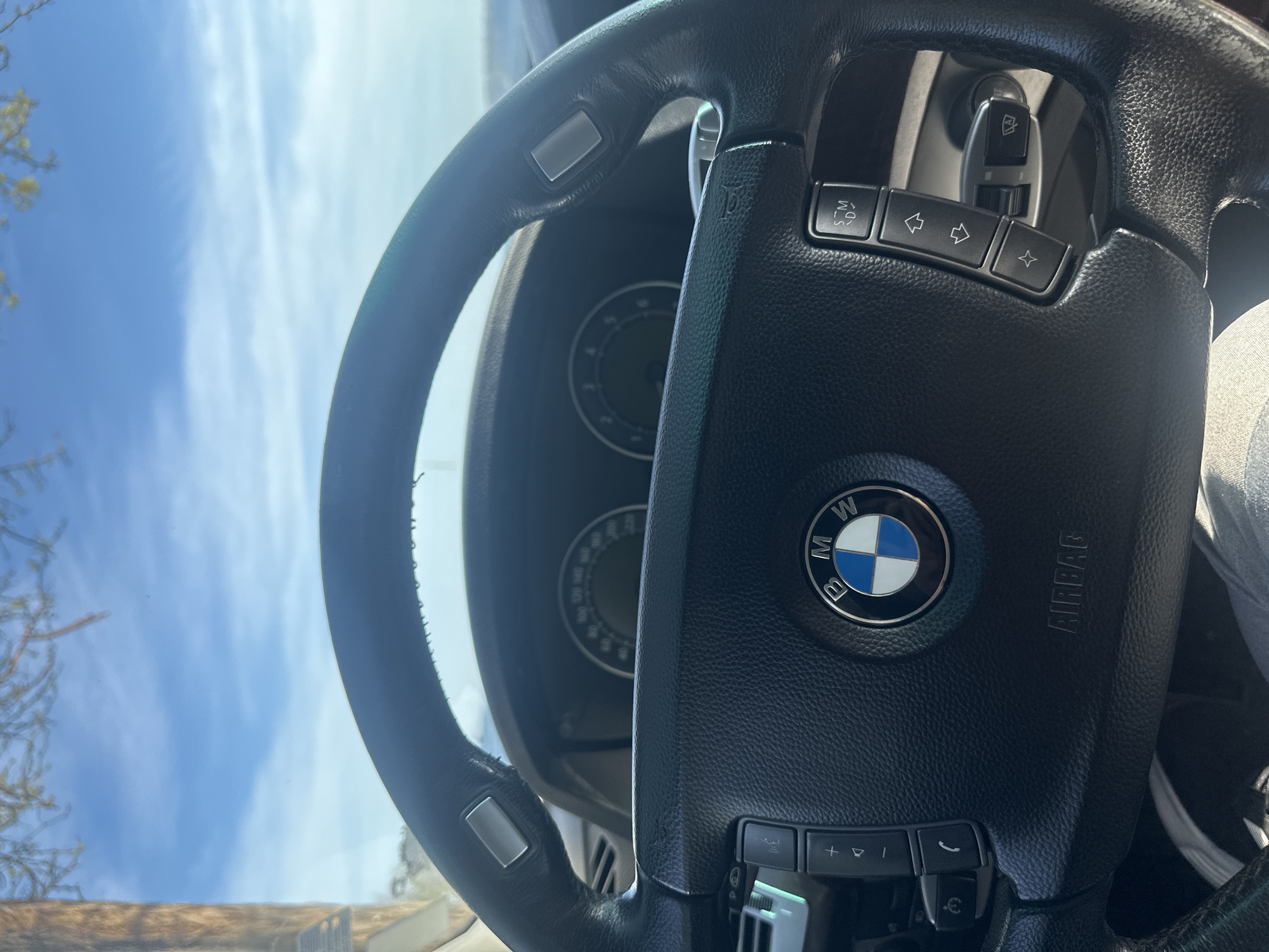 The image displays the interior of a car, focusing primarily on a black leather multi-function steering wheel. The central hub of the steering wheel features the prominent blue and white BMW logo, with the word "AIRBAG" embossed on its right side.
Various control buttons are integrated into the steering wheel spokes. On the left spoke, there are buttons for volume control (+ and - symbols), a phone answer button, and a button with a microphone/speech bubble icon, likely for voice commands or ending calls. The right spoke contains buttons labeled "S/M", left and right arrows (possibly for track or menu navigation), and a star-shaped button, likely a programmable shortcut. Above these, a button with a horn symbol is partially visible. Behind the steering wheel, a stalk with a windshield wiper symbol indicates controls for the wipers.
Behind the steering wheel, a portion of the car's dashboard is visible, showing two circular gauges. Through the windshield, a clear blue sky with some wispy white clouds is seen, indicating a bright, sunny daytime setting. Out-of-focus tree branches with light-colored leaves or flowers are visible on the left side of the exterior view. No individuals are present in the image.