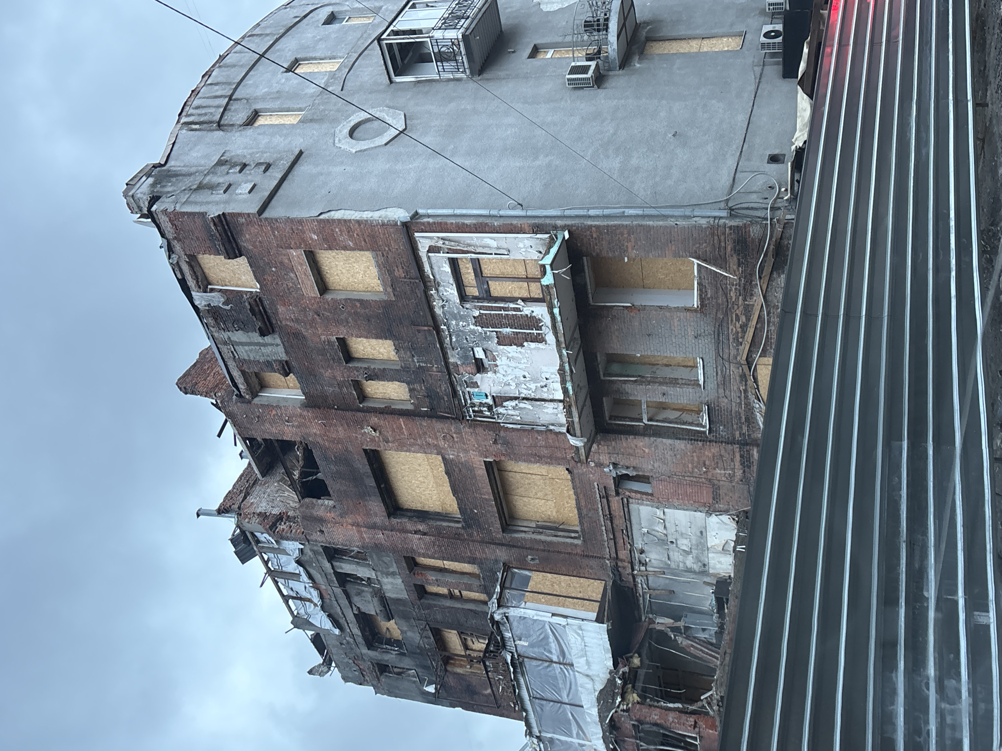 This image depicts a severely damaged multi-story building, likely in Kharkiv, Ukraine, given the provided context. The structure appears to have suffered significant destruction, consistent with conflict or catastrophic event.
The building's lower and middle sections are constructed from red brick, much of which is blackened and crumbling, revealing internal elements and voids where walls or floors once stood. Numerous windows in these sections are boarded up with plywood or similar material. The upper floors of the building feature a smoother, light gray concrete facade, which also shows signs of wear, chipping, and cracks. Architectural details, such as rectangular recesses and a circular design, are visible on the concrete surface. Several small, damaged metal balconies or exterior air conditioning units protrude from the upper facade. Loose wires are strung across the building's exterior.
Parts of the building are visibly collapsing, with debris and exposed structural elements. The upper-left corner of the concrete facade shows significant damage and crumbling. On the right side of the image, a vertical structure composed of corrugated metal panels or a protective barrier stands adjacent to the damaged building. The sky is overcast and gray, suggesting a cloudy day. No people are visible, and there is no discernible text in the image. The scene conveys a sense of desolation and the aftermath of heavy damage.