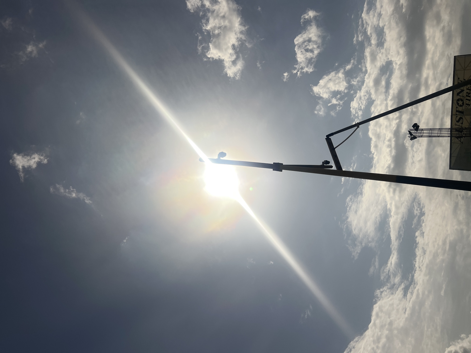 The image captures a sunny sky with scattered clouds. A lamppost stretches across the frame, partially obscuring the sun. A billboard for "STON TIRE" is visible on the right side of the image. The location is in the city of Unknown, Unknown.
