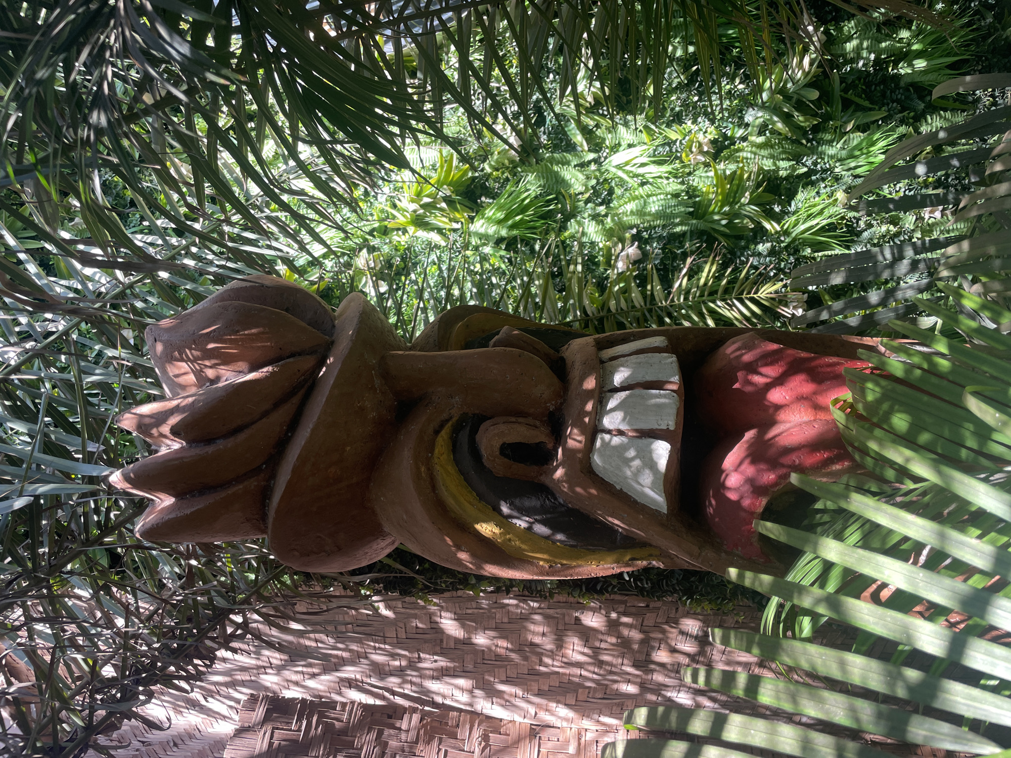 A tiki-style sculpture lies on a woven surface under a canopy of tropical plants. The sculpture is predominantly brown with white teeth and a red section on its right end. The surrounding foliage is lush and green, suggesting a tropical environment. The woven surface below the sculpture is partially obscured by shadows from the overhead plants. While the location of Chopdem, India, is indicated, it is only verifiable by external information and is not directly evident from the visual content of the image itself. There are no visible people.