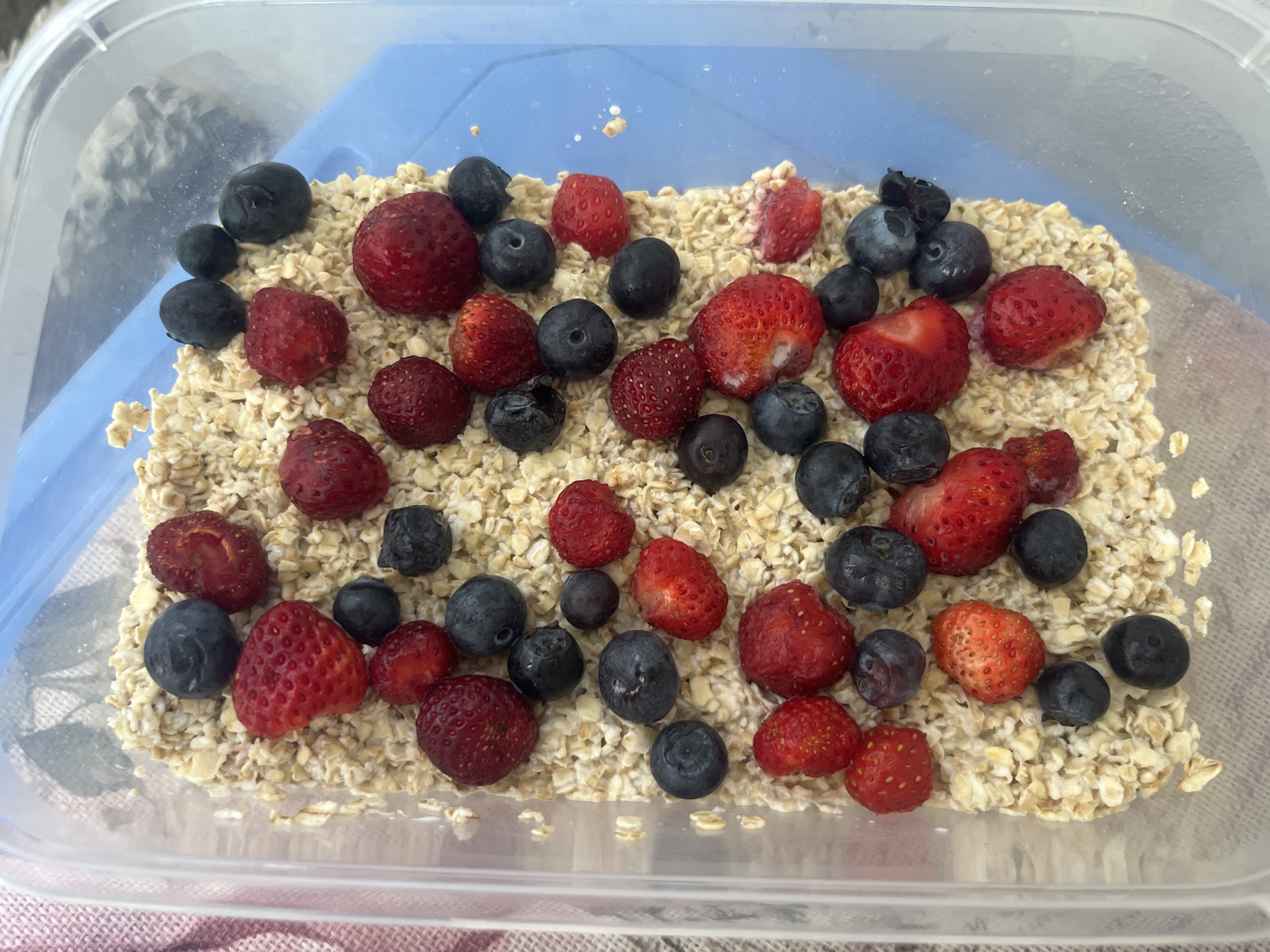 This media file depicts a close-up, overhead shot of a clear plastic container filled with uncooked rolled oats and topped with an assortment of fresh blueberries and strawberries. The berries are scattered generously across the oats, with some partially submerged.
The subjects are the food items: rolled oats, blueberries, and strawberries, arranged within a transparent rectangular container that has a light blue lid visible in the background. The container is resting on a surface with a textured, likely fabric, tablecloth.
The activity taking place is the preparation or display of a breakfast or snack, specifically oatmeal with fruit. There are no people visible, nor any indication of a specific time of day or weather. The setting is likely indoors, given the lighting and the container.
Notable details include the vibrant red of the strawberries, some of which appear to be halved, and the deep blue-black of the blueberries. The texture of the oats is clearly visible. There is no visible text in the image. The overall impression is one of a healthy and appealing meal. The location context of Burgas, Bulgaria, does not offer specific visual cues within the image itself.
