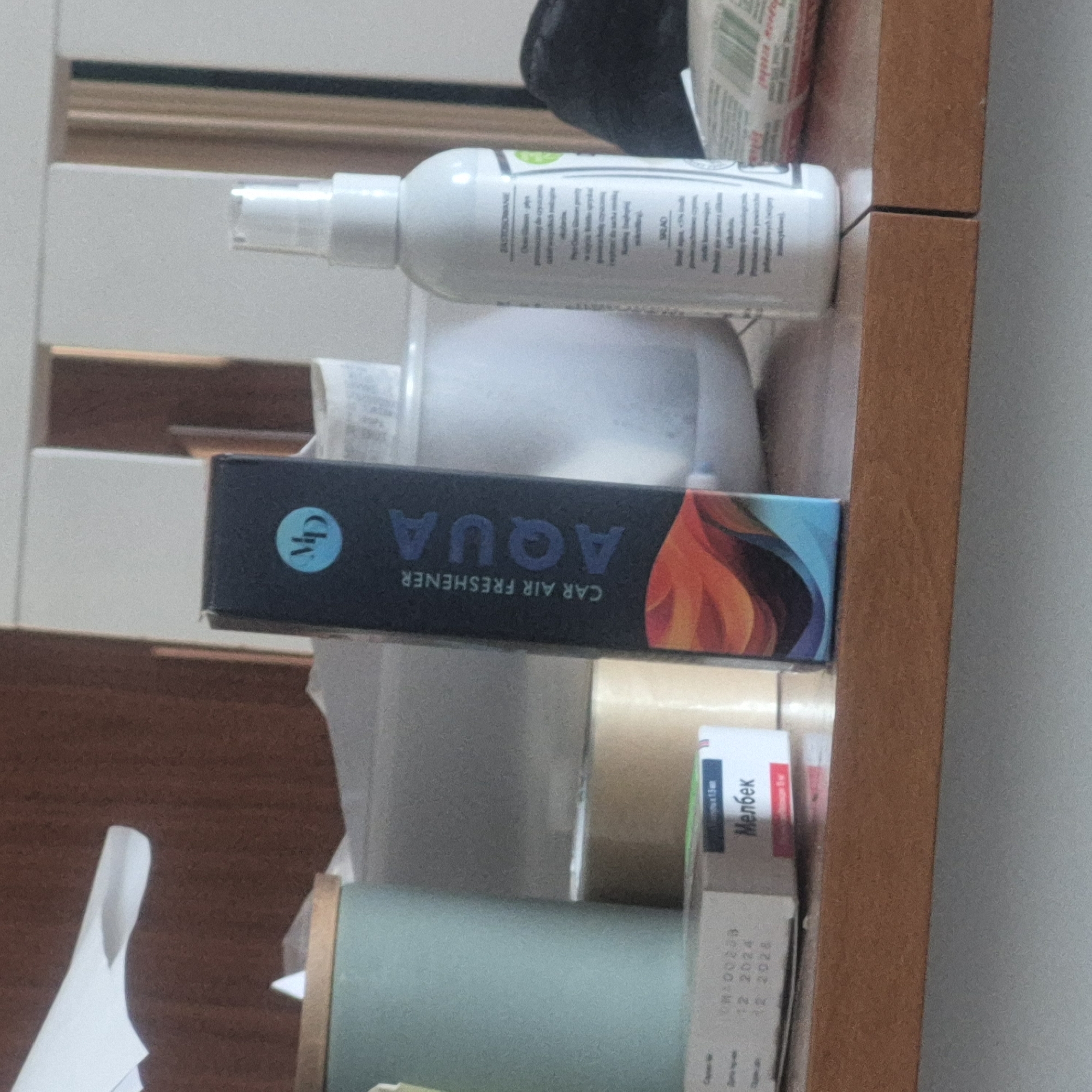 The image shows a cluttered wooden shelf with various items. In the foreground, on the left, is a light blue cylindrical container. To its right, a black box with "AQUA" printed vertically and "CAR AIR FRESHENER" printed horizontally is visible. Next to this, on the right, is a white spray bottle with a clear cap. Behind these items, parts of other objects and a white shelf are discernible. In the bottom left corner, a small white box with Russian text and dates is present. The overall scene appears to be an indoor environment, possibly a room or a storage area. The image was taken in Kępno, Poland.