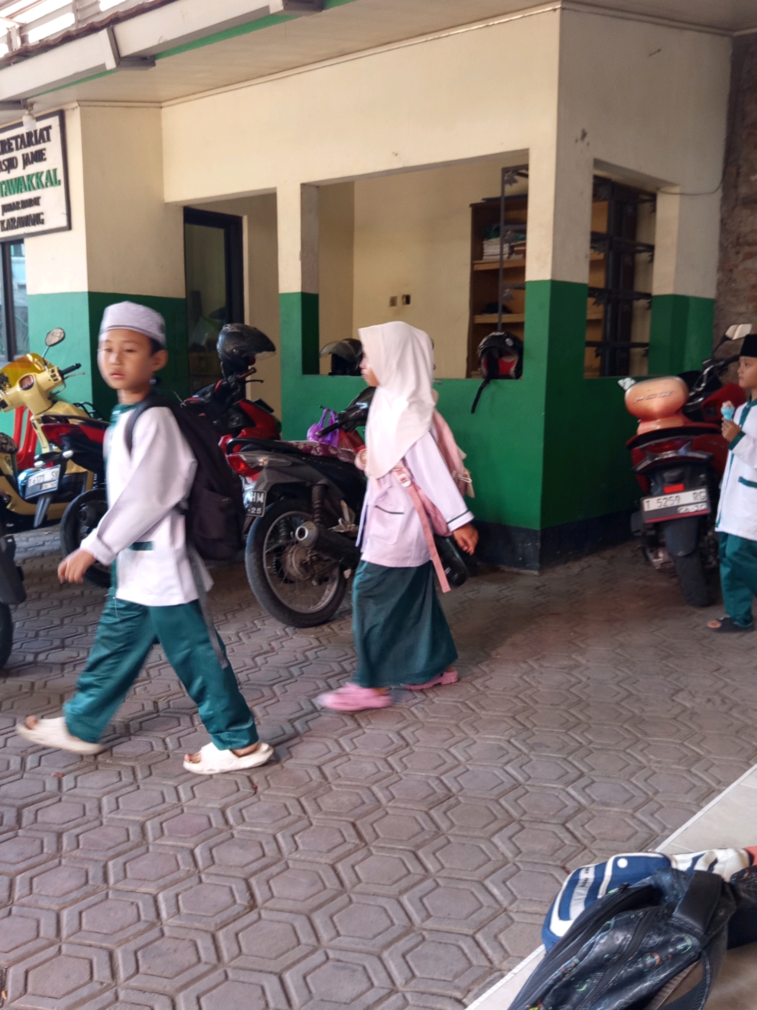In front of a building with a sign that reads "SEKRETARIAT MASJID JAMIE TAWAKKAL" in Karawang, Indonesia, two children wearing Islamic school uniforms are walking on a patterned concrete pathway. The child on the left, a boy wearing a white long-sleeved shirt, green pants, and a white cap, carries a black backpack and white sandals. The child on the right, a girl, wears a white headscarf, a light pink long-sleeved shirt, and a dark green skirt, with pink sandals. Several motorcycles are parked behind the children. The building has a white and green exterior with a covered walkway.