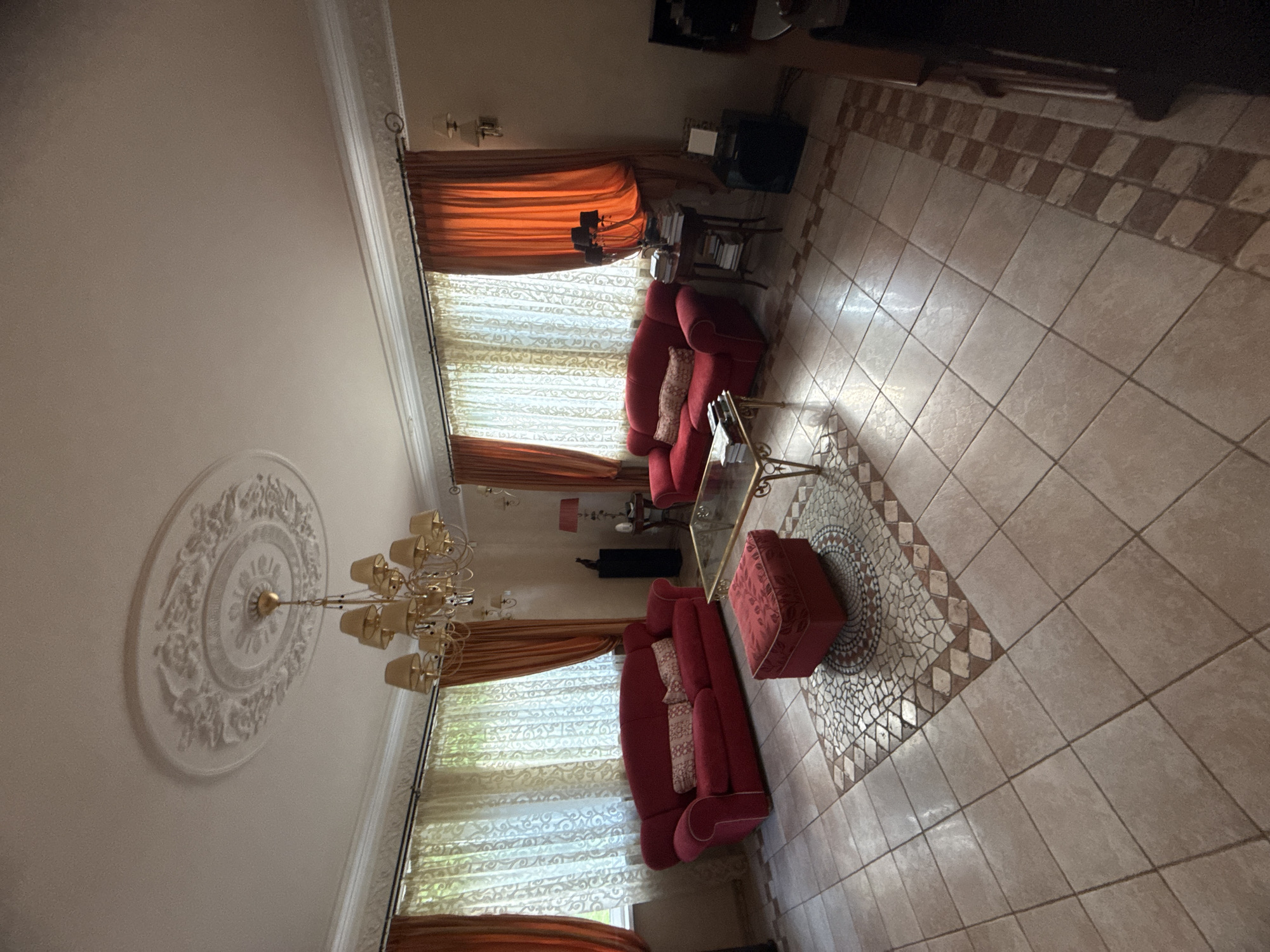 The image depicts a living room in a dwelling, possibly in Yukki, Russia. The room is furnished with two red sofas, a matching ottoman, and a glass-topped coffee table. A large chandelier hangs from a decorative ceiling medallion, and the windows are dressed with sheer and orange drapes. The floor is tiled with a mosaic inlay pattern in the center. The scene appears to be unoccupied.