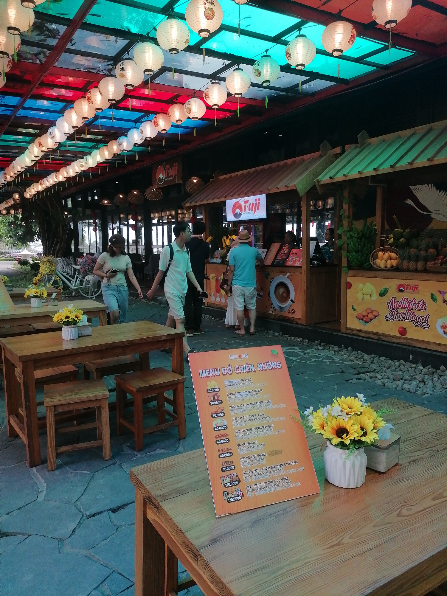 The image shows an outdoor food area, likely a restaurant, in Hong Gai Ward, Vietnam. A row of food stalls is visible, including one with a "Fuji" sign. A couple walks past the stalls, while other people are at the stalls, possibly ordering food. Tables and chairs are set up for dining, with a menu board on one table. The area is decorated with hanging lanterns. The ground is paved with irregular stones. There are some flowers in pots on the tables.