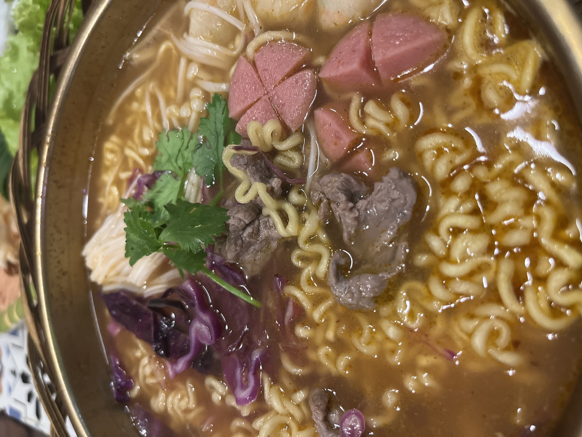 A close-up shot shows a bowl of Vietnamese noodle soup. The soup is a rich brown broth filled with wavy yellow noodles, thin white noodles, slices of pink processed meat, pieces of thinly sliced beef, and chunks of purple cabbage. A sprig of green cilantro garnishes the top. The bowl has a golden rim and appears to be part of a larger, possibly communal, dining setup, with blurred greenery visible in the background, suggesting an outdoor or restaurant setting. The lighting is warm and direct, highlighting the textures and colors of the food. The scene is static and focuses solely on the food.