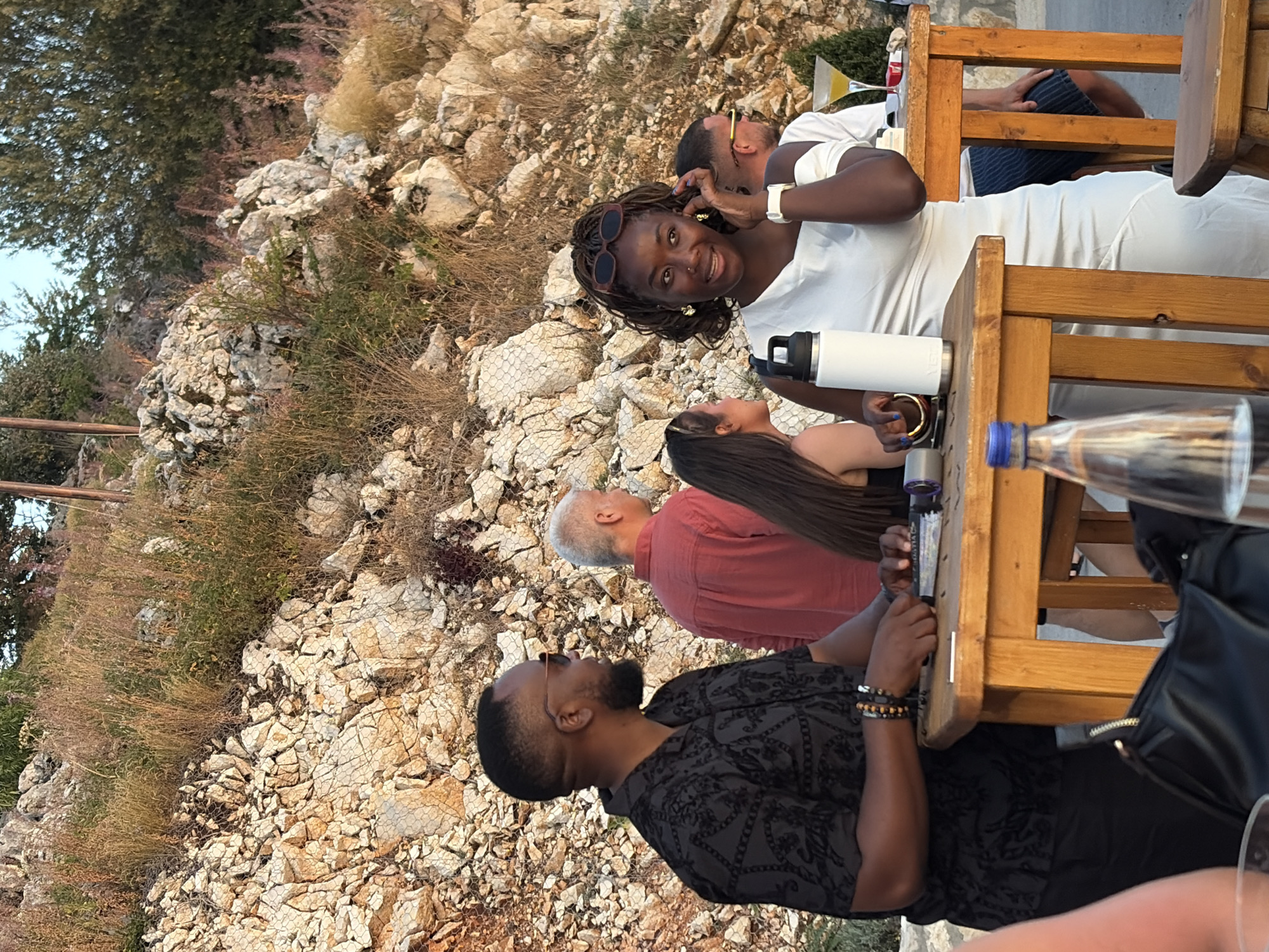 In Kuk, Montenegro, a group of people are gathered at an outdoor seating area, likely a restaurant or bar, situated on a rocky hillside with sparse vegetation. A Black man in a patterned dark shirt and sunglasses is on the left, facing right, with his hands resting on a wooden table. He appears to be interacting with something on the table. To his right, a Black woman in a white off-the-shoulder dress and sunglasses is holding her phone to her ear and looking to her right, engaged in a conversation. A white water bottle is on the table in front of her. Behind the man, a fair-skinned man in a red shirt is also visible. Further to the right, another man in a white shirt and sunglasses is partially obscured. The setting sun casts a warm glow on the scene, highlighting the rocky terrain and the relaxed atmosphere of the gathering.