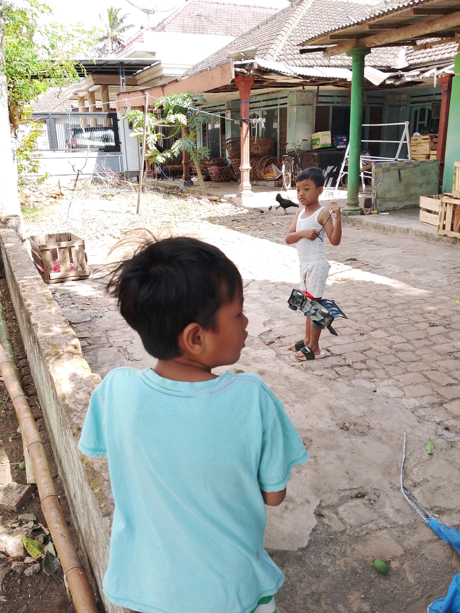 In an outdoor, dusty courtyard, two young boys are observed. The boy in the foreground, facing away from the camera, wears a light blue t-shirt. His dark hair is disheveled. The second boy, in the mid-ground and facing the camera, wears a white tank top and patterned shorts. He holds a black and silver toy car attached to a string. Several chickens are present on the ground. The background reveals a single-story building with a tiled roof, supported by green and red pillars. Wooden crates and a bicycle are visible near the building. The ground is paved with cobblestones. The overall setting suggests a residential area in Karangsari, Indonesia.