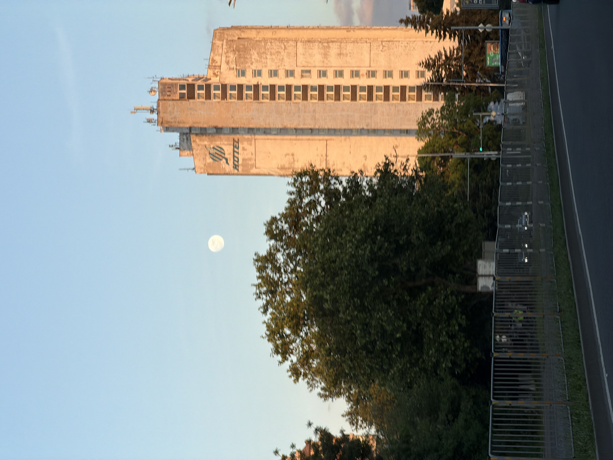 The image depicts the Hotel Bulgaria in Burgas, Bulgaria. The tall, rectangular building is visible in the background, featuring a faded "HOTEL" sign on its side. In the foreground, there are trees and a metal fence running along the edge of a road. A full moon is visible in the sky to the left of the hotel. The image is taken during daylight hours, with a clear blue sky. The hotel's facade is weathered, suggesting age and possible lack of recent maintenance. The surrounding environment is a mix of urban development and greenery. A green traffic light is seen behind the trees.