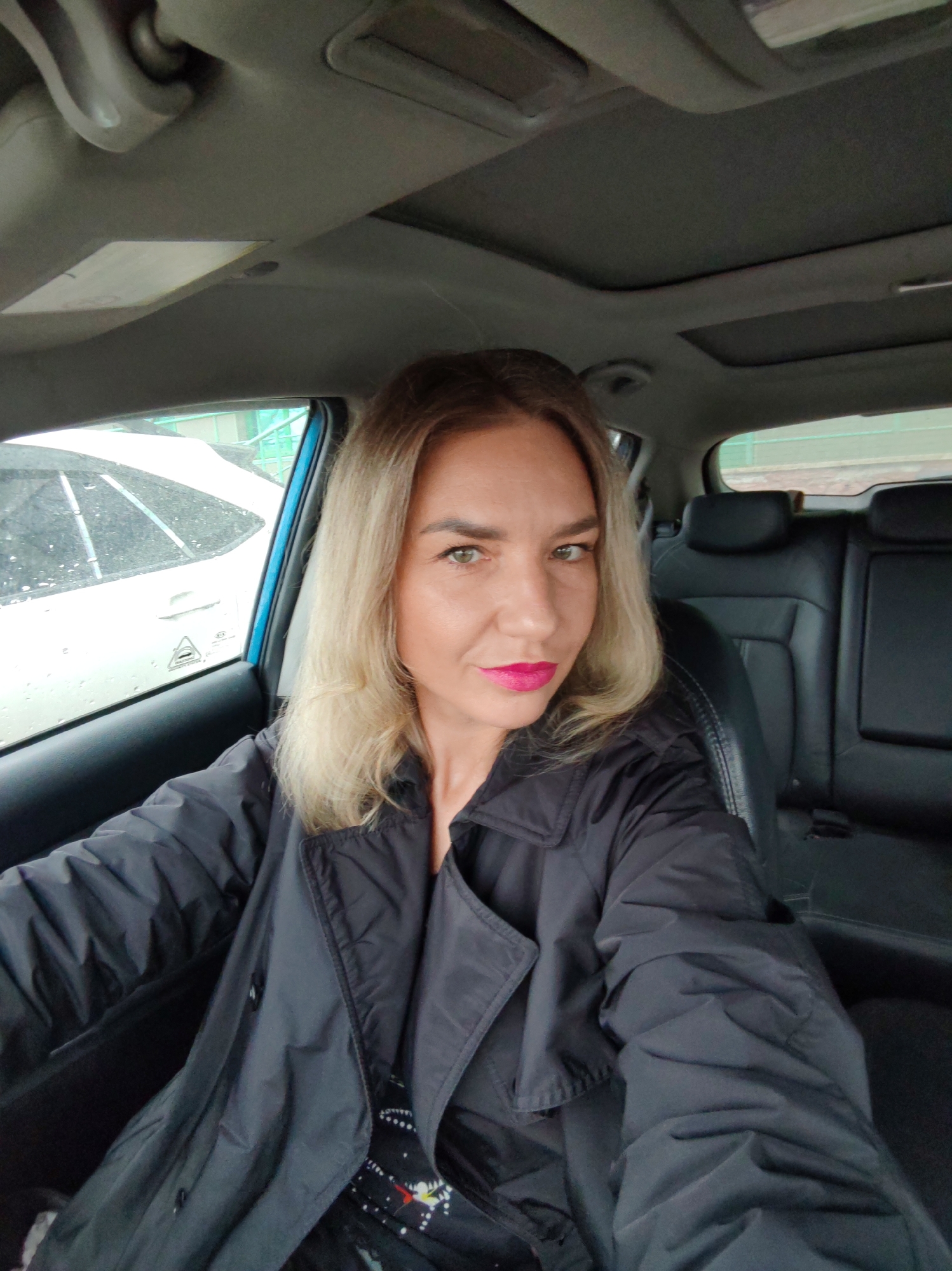 A woman is seated in the driver's seat of a car, facing the camera. She has blonde hair and is wearing a dark coat with visible buttons. The car has a sunroof, and there is another vehicle visible through the driver's side window. The car's interior is primarily dark. The location in Petropavlovsk-Kamchatsky, Russia is not determinable from the visual data alone.