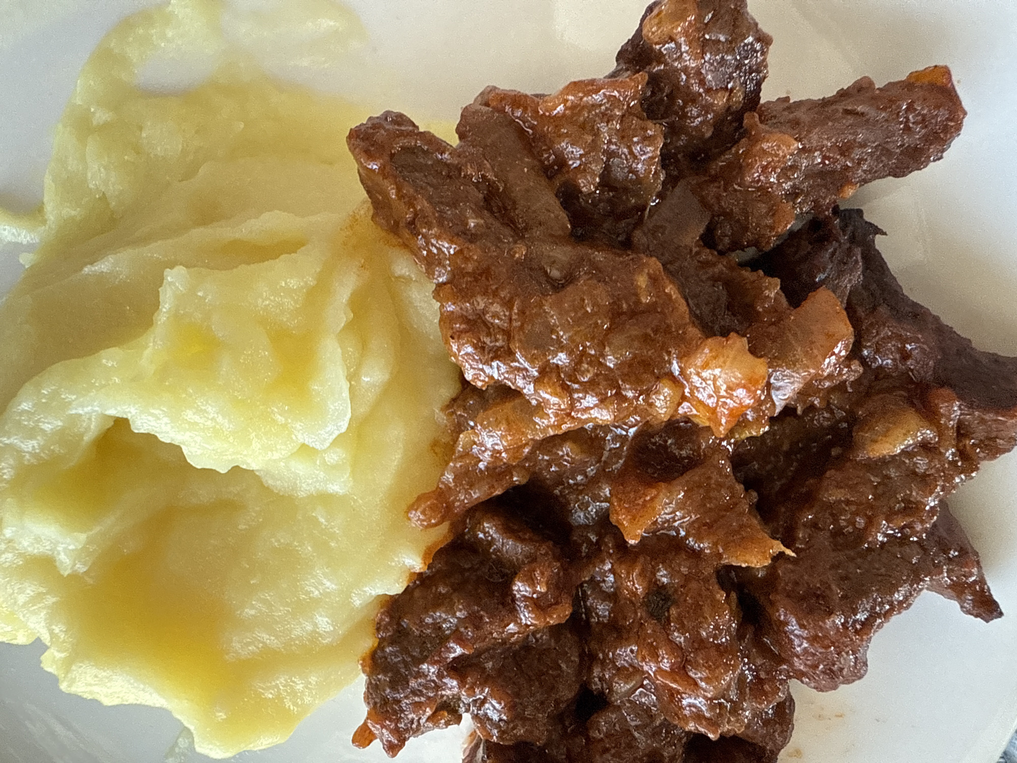 An overhead, close-up shot captures a serving of food on a white plate. The plate is divided almost vertically by two distinct food items.
On the left side of the frame is a portion of mashed potatoes. They are a pale yellow color with a smooth, slightly lumpy texture, indicating a creamy consistency. The surface shows some gentle undulations and shadows, suggesting its soft form.
Occupying the right side of the frame is a generous serving of dark brown, glossy meat. The meat appears to be stewed or braised, cut into irregular strips and chunks. It has a rich, deep brown hue, and the surface is shiny, likely from a savory sauce or gravy that coats it. The individual pieces of meat are piled together, creating a textured mound.
The composition highlights the two main components of the meal, with the white plate providing a neutral background. There are no people visible, nor any specific activity beyond the presentation of the meal. No text is visible within the image. While the provided context indicates the location is Votkinsk, Russia, there are no visual cues within the image itself to confirm this. The lighting is bright and even, suggesting an indoor setting but giving no indication of the time of day or weather.