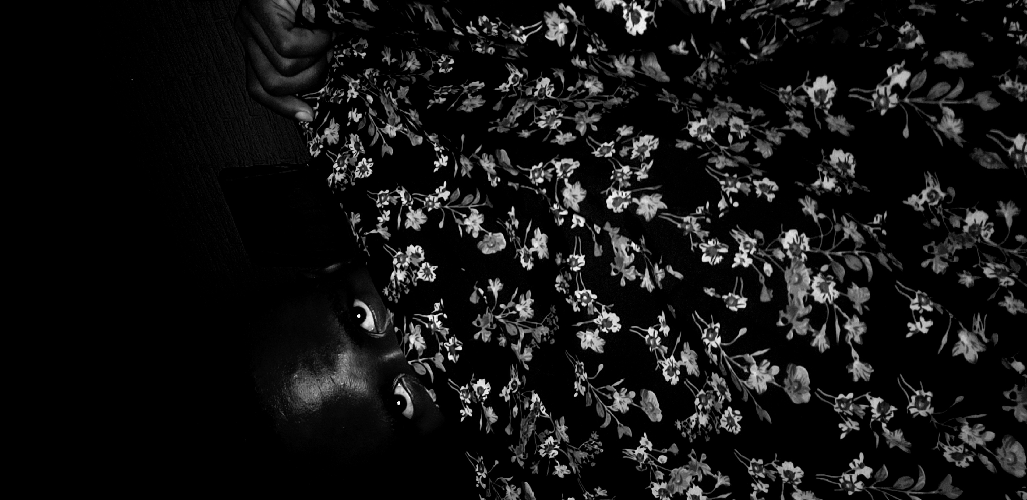 A black and white image, likely a photograph, depicts a person partially obscured by a floral patterned fabric. The person's face, specifically the forehead, nose, and eyes are visible on the lower left side of the image. The subject's eyes are wide and gazing upwards. A hand, seemingly belonging to the same individual, holds a section of the floral fabric near the upper left portion of the frame. The floral pattern consists of small, light-colored flowers with accompanying leaves, set against a dark background. The remainder of the image is dark, obscuring any clear sense of environment or location. No specific indicators of the city of Narayi, Nigeria, are present in the visible details of the image.