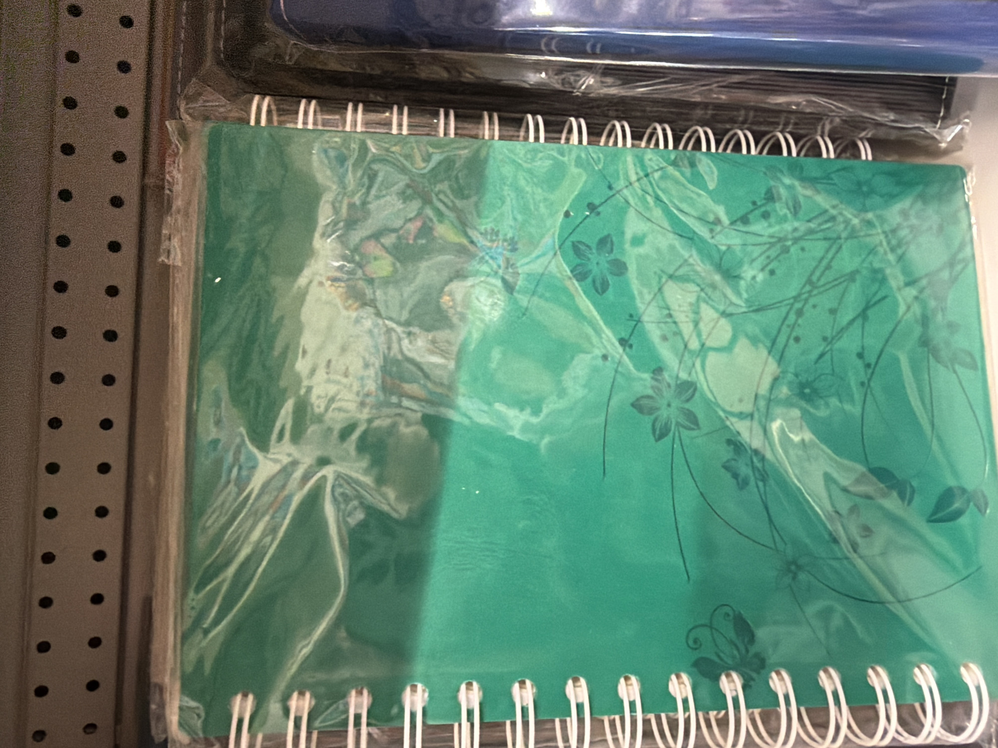A close-up, eye-level, indoor shot shows the side of a spiral-bound notebook with a green cover, encased in clear plastic. The notebook is lying flat on a shelf, with a portion of another notebook, likely a dark blue one, visible to its right. The notebook cover features a dark floral and butterfly design. The background consists of a grey, perforated shelf. The location is a retail store shelf in Pazardzhik, Bulgaria.