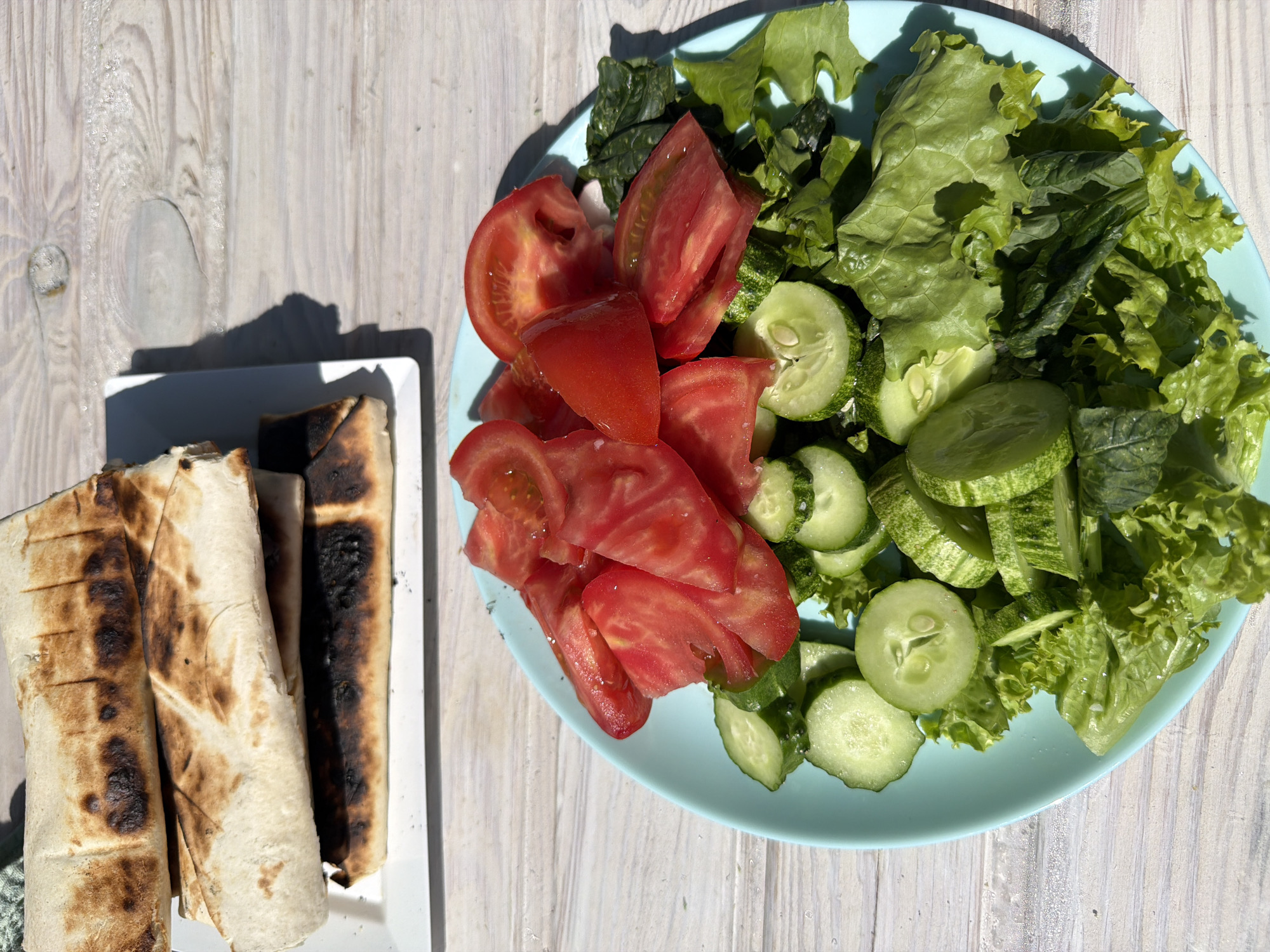 A high-angle, outdoor shot shows a plate of food on a light-colored wooden surface. To the left, on a white plate, are four grilled wraps, some with burn marks. To the right, a blue plate holds a salad with sliced tomatoes, cucumbers, and leafy greens. The photo does not provide any information about the location in the city of Chaikovsky, Russia or any information about people, actions, or interactions.