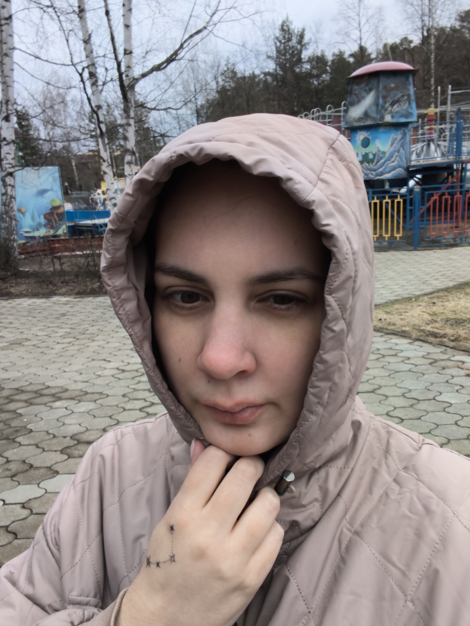 A close-up, eye-level portrait features a woman wearing a light pink or beige quilted hooded jacket, with the hood pulled up. Her gaze is directed downwards, giving her expression a neutral, perhaps pensive or slightly tired appearance. Her right hand is gently positioned near her chin, revealing a small tattoo on the back of her hand, resembling a constellation of connected stars.
The scene is set outdoors in what appears to be a park or playground in Chaikovsky, Russia. The ground is paved with interlocking concrete blocks forming a patterned surface. In the background, several tall, slender trees with white bark (likely birches) are visible, mostly bare of leaves, suggesting either early spring or late autumn. Beyond the trees, colorful playground structures are evident, including a blue and red fenced area and a multi-tiered play structure with painted panels depicting scenes like underwater life and stylized landscapes. A large blue board with a detailed underwater mural is prominent on the left side of the background. The lighting is soft and diffused, characteristic of an overcast day, contributing to a muted color palette throughout the image. There is no legible text visible within the frame.