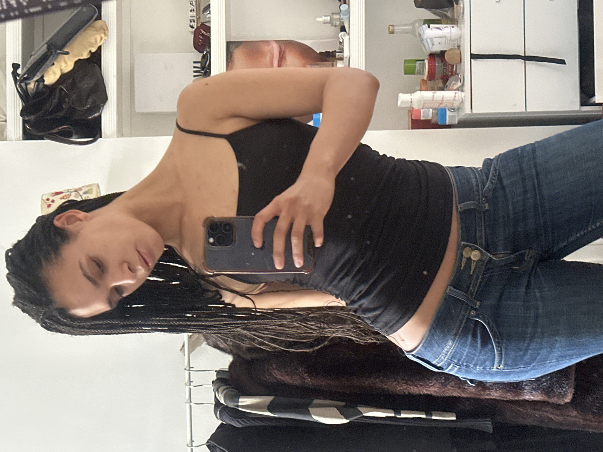 A young woman with dark braided hair is taking a selfie in a bathroom mirror. She is wearing a black tank top and blue jeans. The bathroom has white cabinets and various toiletries on the shelves. There are clothes hanging below the mirror. The location is not identifiable as being in London, United Kingdom from the information provided in this image.