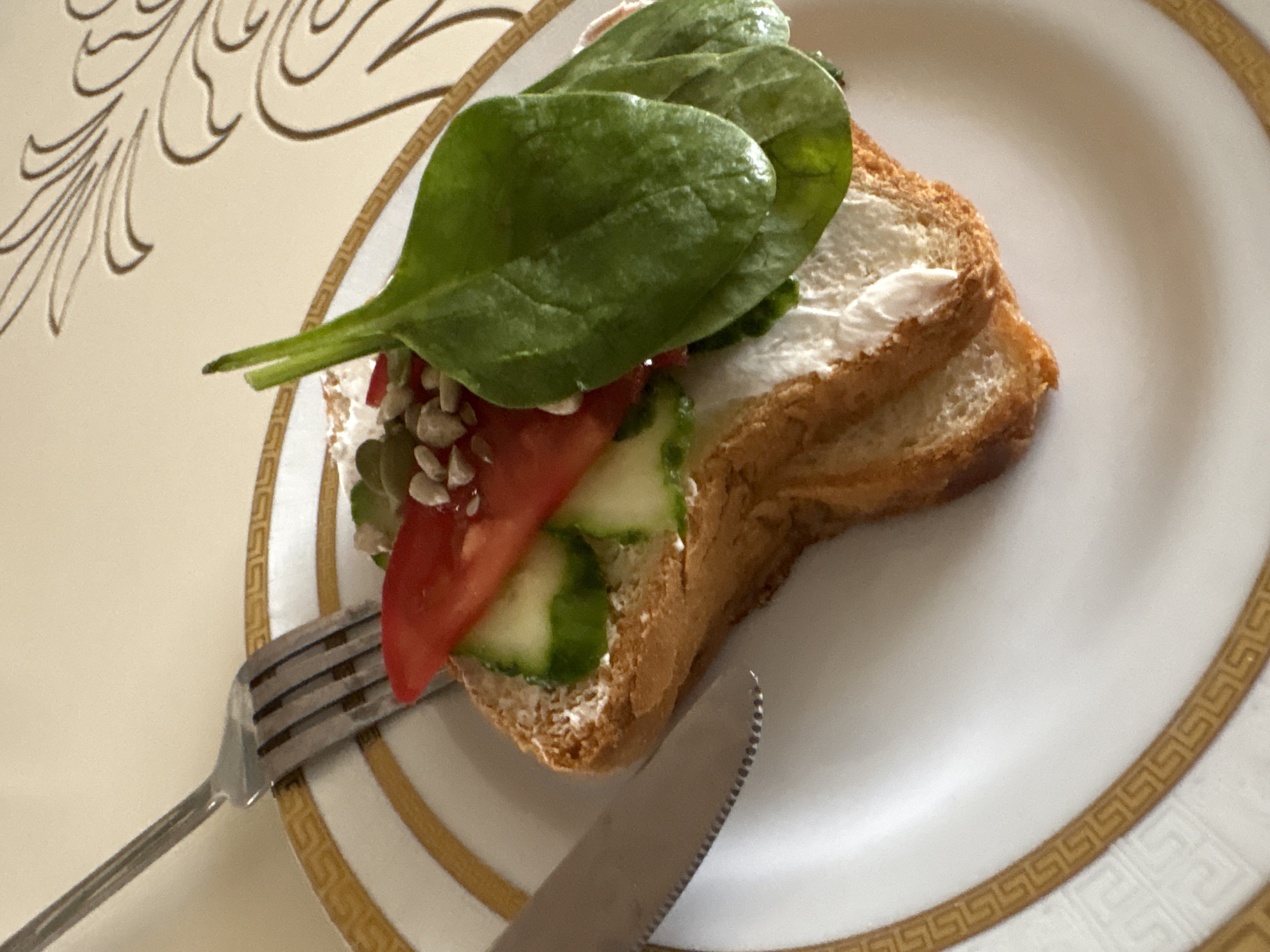 A close-up, high-angle shot shows a slice of toast topped with cream cheese, cucumber slices, a tomato slice, and spinach leaves on a white plate with a gold Greek key pattern border. A fork and knife are placed to the left of the plate, and a decorative embossed pattern is visible on the table in the background. The image is a photograph taken in Georgiyevsk, Russia.