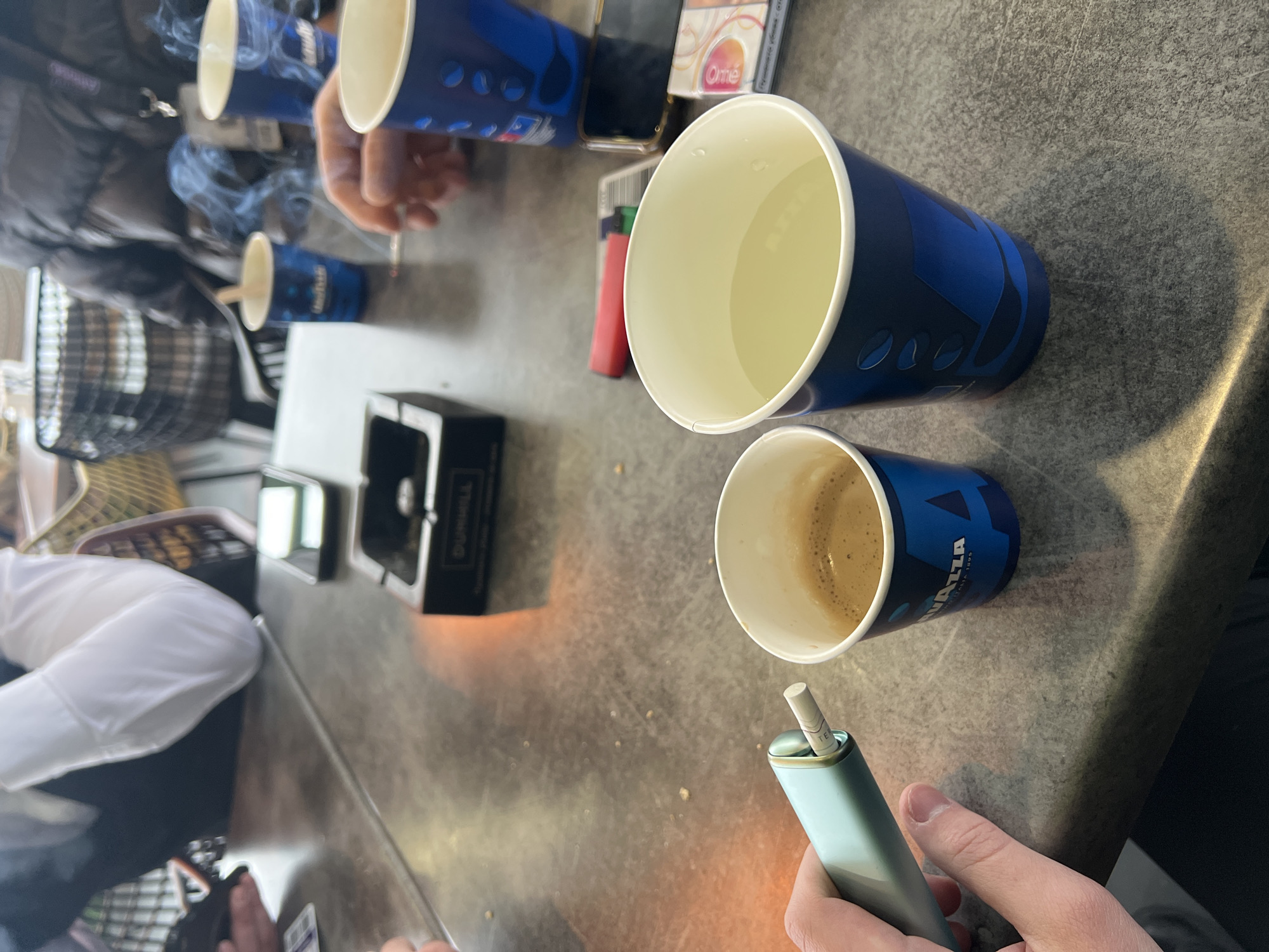 The image shows a table scene, likely in a café or casual setting. Several blue "LAVAZZA" branded paper cups are present, some containing liquids, including coffee. Visible on the table are an ashtray, a lighter, a pack of cigarettes, and a cell phone. Smoke is visible in the air, indicating recent cigarette use. A person is holding a light-blue device with a cigarette inserted. Based on the artifacts present, the location may be a public area or café in Sofia, Bulgaria, where Lavazza coffee and cigarettes are commonly available. The presence of multiple people and objects indicates a gathering or meeting is taking place.