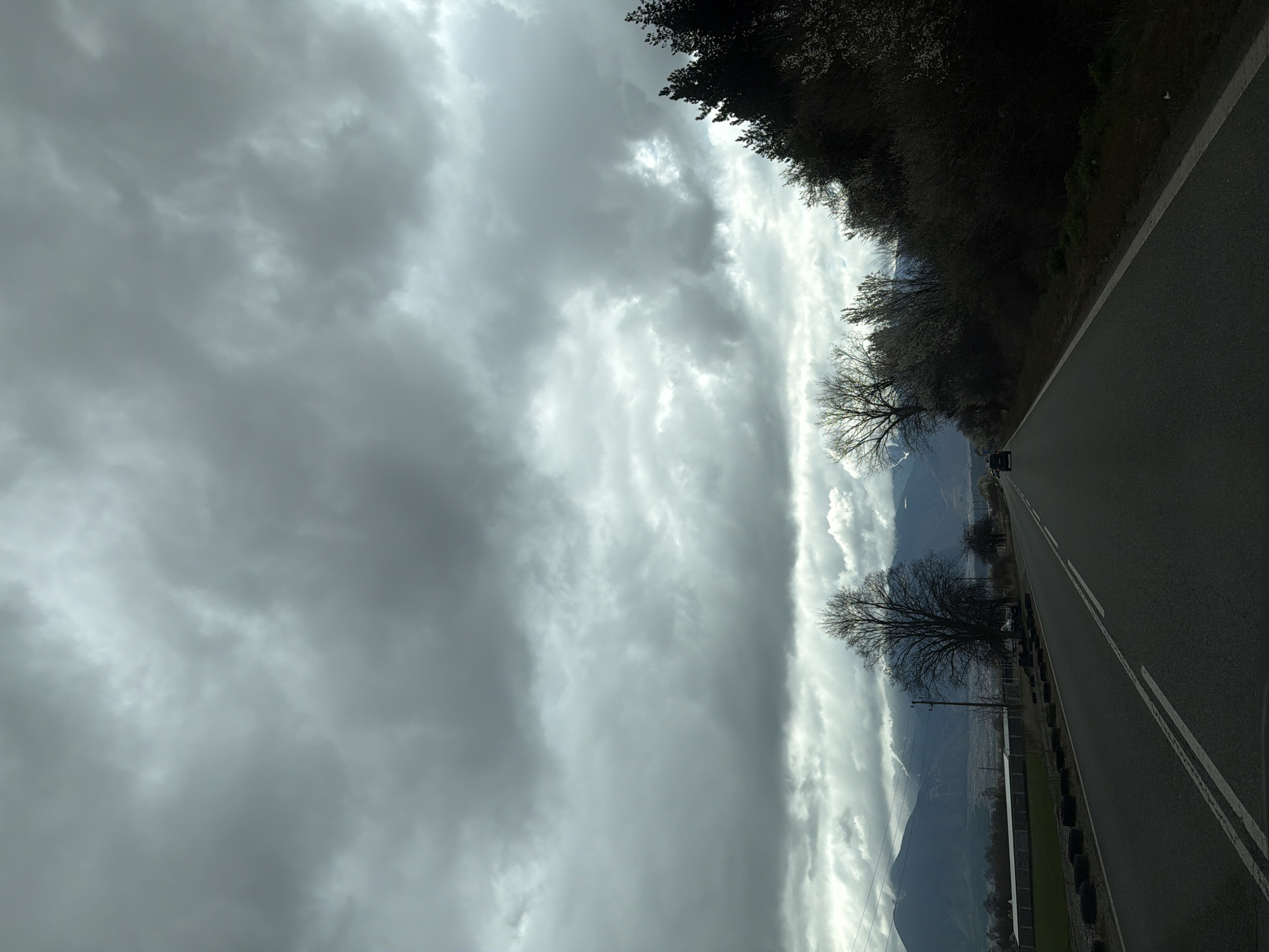 The image shows a cloudy sky above a road, with trees lining one side. A vehicle is seen on the road in the distance. There is a mountain range visible under the clouds. The location and city is not apparent from this image.