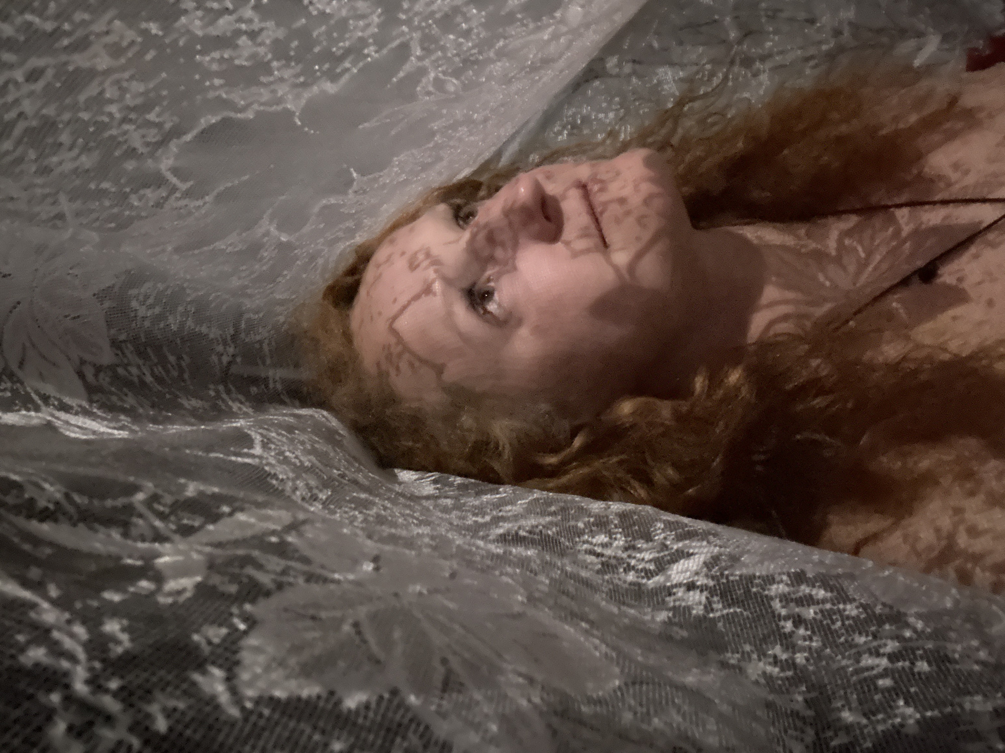 A medium shot presents a person with reddish-brown curly hair lying beneath a semi-transparent, lace-like fabric. The fabric is draped over the person, creating shadows and patterns on their face. Lines, seemingly from the fabric's pattern, are visible on the face. The person is looking upward. A thin, dark cord is visible near the neck, possibly a necklace. The background is indistinct, suggesting the person is indoors. The scene does not provide sufficient information to determine a location in Chaikovsky, Russia, or any information about actions, interactions, and the environment.