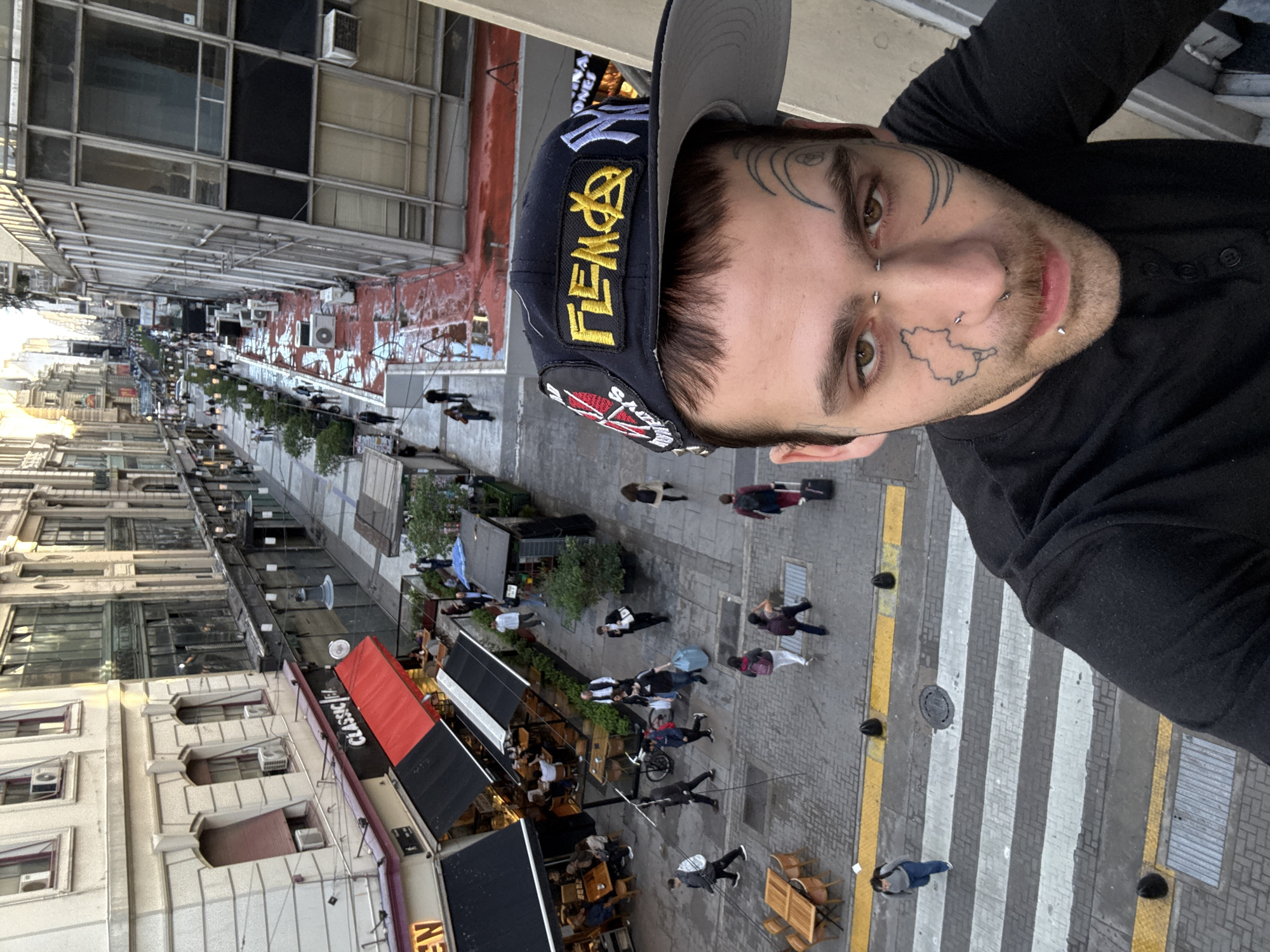 A man takes a high-angle selfie from an elevated position, overlooking a bustling city street in Buenos Aires, Argentina. The man, in the foreground, has a neutral expression, light brown eyes, and several facial piercings including a nose ring, philtrum piercing, and lower lip piercings. His face is adorned with distinctive tattoos: geometric patterns around his eyes and a map-like outline on his right cheek. He wears a black long-sleeved shirt and a grey and navy blue baseball cap. The cap features a prominent yellow patch with stylized text resembling "FEM$€" and a New York Yankees logo.
The background shows a lively urban street lined with multi-story buildings of varying architectural styles, some with traditional elements and others with modern, large windows. Air conditioning units are visible on several facades. The street level features a pedestrian area with paving, small trees, and outdoor seating for restaurants. Numerous pedestrians are seen walking, some carrying bags or a suitcase, and one appears to be with a bicycle. A prominent zebra crossing with white and yellow markings is visible on the street.
The scene is illuminated by soft, natural light, suggesting late afternoon or early evening. The weather appears clear. Visible text includes "CLASSIC" and a partially visible "NEW" on a red and black awning over a restaurant or shop. The overall atmosphere is that of a vibrant, active city environment.