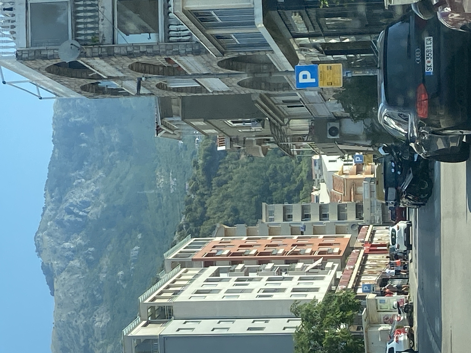 The image shows a street scene in Budva, Montenegro. In the foreground, several cars are parked and driving on a paved road. A black Audi is prominently featured on the right side of the frame. To the left, a white car and a motorcycle are visible.
The street is lined with multi-story buildings on both sides. On the left, a tall, modern building with a white facade and balconies dominates the view. Adjacent to it is a building with a reddish-orange facade. On the right, older buildings with stone accents and arched windows are present, some with balconies. A blue parking sign is mounted on a pole in front of these buildings.
In the background, a lush green mountainside rises steeply. The sky is clear and blue. A few people are seen walking on the sidewalk in front of the buildings. The overall impression is of a busy urban area nestled against a natural landscape.