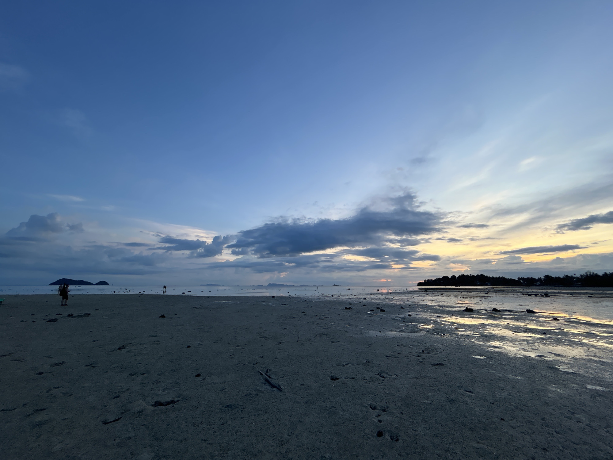 An outdoor, wide-angle, eye-level shot captures a serene beach scene at dusk on Koh Phangan, Thailand. The sky is a gradient of deep blue at the top, transitioning to lighter blues and soft yellows near the horizon, indicating the sun has recently set. Scattered clouds, some dark and others tinged with the fading light, dot the sky.
The foreground is dominated by a damp, sandy beach, reflecting the sky in shallow pools of water. Small rocks and debris are scattered across the sand. In the mid-ground, a calm sea stretches out to the horizon. A few distant islands are visible as dark silhouettes against the brighter sky.
There are a few human figures present on the beach. To the left, a person stands near the water's edge. Further into the distance, a small group of people are walking along the shoreline. The overall atmosphere is tranquil and peaceful, with the fading light creating a soft and muted color palette.