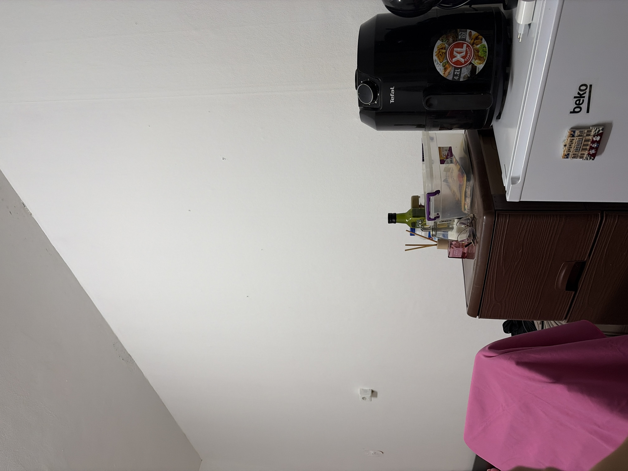 The image depicts a cluttered interior space. A black air fryer labeled "Tefal" and a white "Beko" refrigerator are positioned atop a brown wooden cabinet with drawers. Various items are stacked on the cabinet, including a clear plastic container, a green bottle, and other miscellaneous objects. A pink fabric item is draped over the edge of the cabinet, extending downwards. The walls are white. The location appears to be an indoor space, likely a kitchen or storage area. There are no people visible in the image. It is impossible to ascertain whether it is in Sofia, Bulgaria.