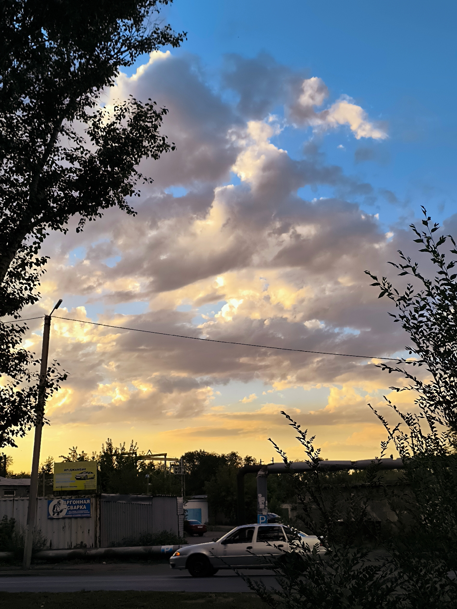 The image depicts a street scene in the city of Qarağandı, Kazakhstan, during sunset. The sky is filled with dramatic clouds, tinged with yellow and orange from the setting sun, contrasting with patches of blue sky. In the foreground, silhouetted trees frame the view on the left and right. A street lamp stands on the left side of the frame, with a power line stretching horizontally across the middle ground. Below the sky, a street runs horizontally. A light-colored car is visible driving on the street. In the background, there are buildings, including a corrugated metal structure with signage, and various trees. One sign, in yellow, features an image of a car. Another sign, with blue and white lettering, advertises "ARGON WELDING." The overall impression is a typical urban landscape at dusk, with the dynamic sky as the focal point.