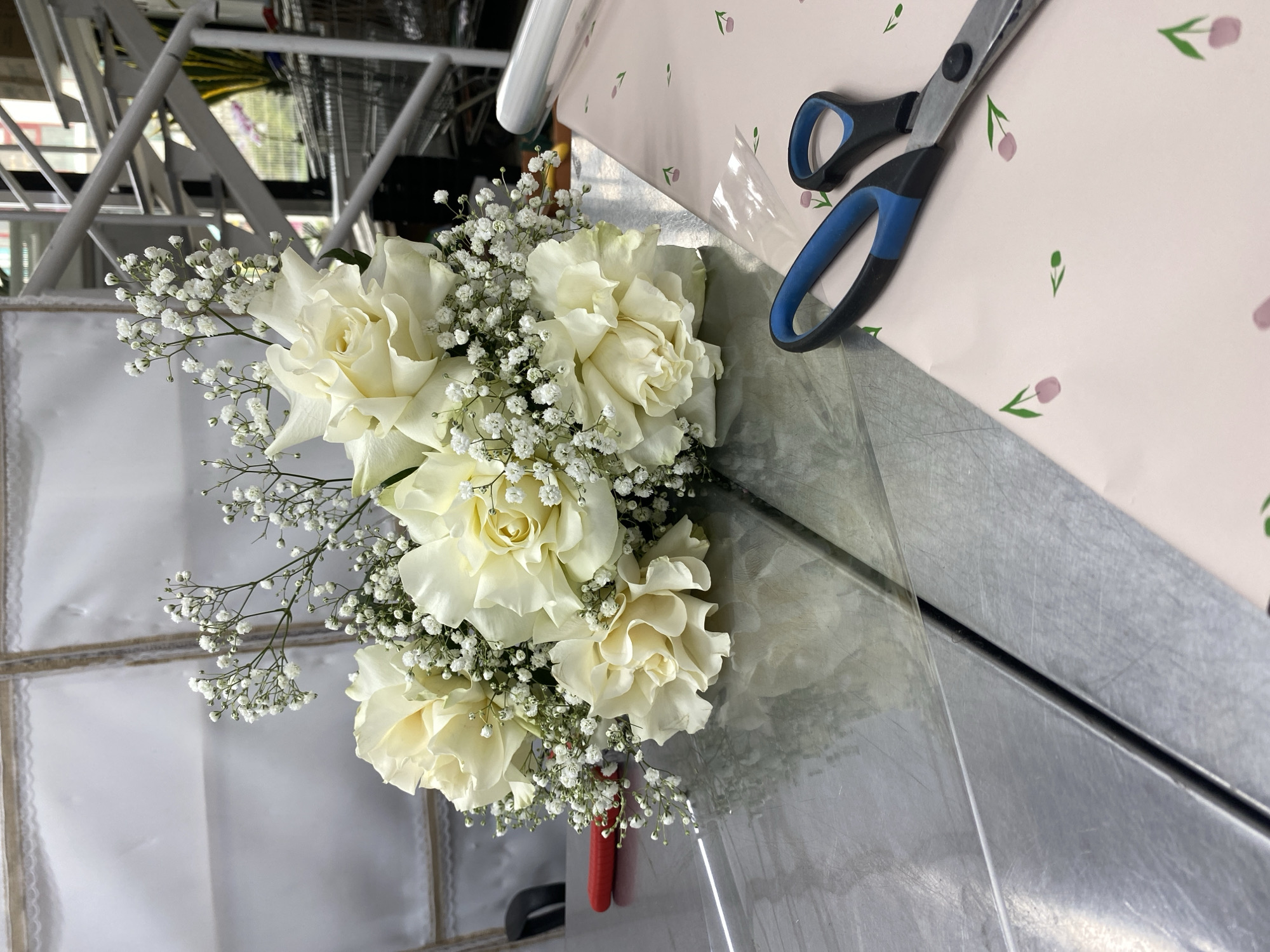 A close-up shot features a bouquet of white roses and baby's breath flowers on a metallic surface. To the right of the bouquet lies a pair of scissors with blue and black handles, positioned on a pink surface with a small flower pattern. The bouquet is wrapped in clear plastic, and a red ribbon is visible at the base of the stems. In the background, a white metal structure suggests a workshop or studio setting. The location is not discernible from the visual information provided, and a statement about Yekaterinburg, Russia cannot be made.