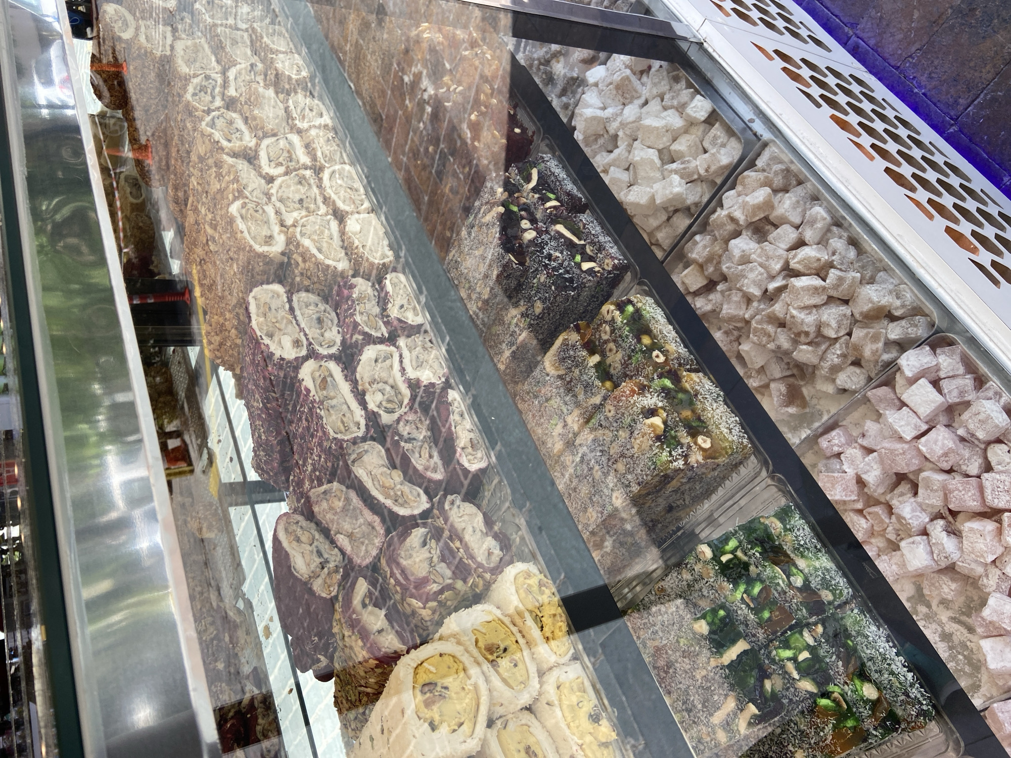 The image captures a display case filled with various types of Turkish delight. The case is likely located in a shop in Antalya, Turkey, given the traditional Turkish sweets. The display features different flavors and presentations, including rolled Turkish delight with nuts, squares covered in coconut flakes and nuts, and cubes dusted with powdered sugar. The sweets are arranged in rows, showcasing the variety of available options. The image is taken from an elevated angle, looking down into the display case. The lighting is bright, highlighting the colors and textures of the Turkish delights. The overall impression is of a tempting selection of traditional Turkish sweets for sale.