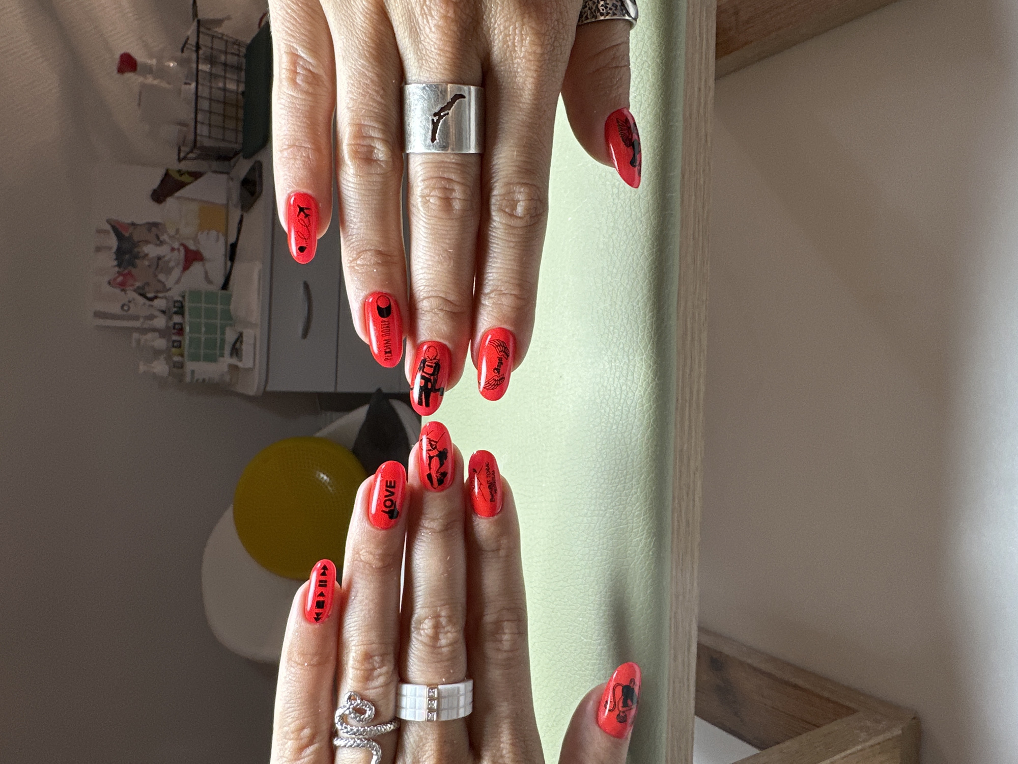 This is a close-up, eye-level, indoor, medium shot of two manicured hands with bright red nails, each adorned with black decals. The hands are positioned over a light green surface, which appears to be the top of a table or stand, supported by wooden legs visible at the bottom of the frame.
The left hand has several rings: a silver coiled ring on the index finger, and a white rectangular ring with three small square gemstones on the ring finger. The red nails on this hand feature black decals: the thumb has two small figures, the index finger has play and skip buttons, the middle finger has the word "LOVE" in black text, and the ring finger has a black decal of two figures dancing or interacting. The pinky finger has a black decal of what appears to be a winged figure.
The right hand also has red nails with black decals. The index finger has a black decal of two figures in a similar pose to the left hand's middle finger. The middle finger has a black decal of what looks like an airplane, and the ring finger has a black decal of what appears to be a winged figure, similar to the left pinky. The thumb has a black decal of what might be a flower. On the ring finger of the right hand is a silver ring with a cutout silhouette of what appears to be the outline of the Crimea peninsula.
The background is out of focus, showing a shelf with various items including bottles, a basket, and a framed picture of a cat. The lighting suggests an indoor setting, possibly a room or a studio. The image does not provide any information about the city of Vladimir, Russia.