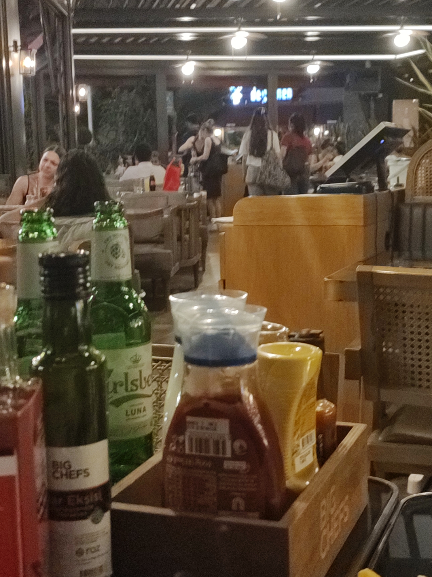 The image depicts a dimly lit, indoor dining area of a restaurant, possibly in Kadıköy, Turkey, given the presence of a Turkish brand of beer. In the foreground, several bottles are visible, including two green glass beer bottles, one with a white label that appears to say "Carlsberg LUNA", and another dark green bottle with a white label that reads "BIG CHEFS EKSI". A ketchup bottle and a yellow mustard bottle are also present. In the background, blurred figures of people are seated at tables, some interacting. The overall ambiance suggests a casual dining setting with ambient lighting from overhead fixtures.