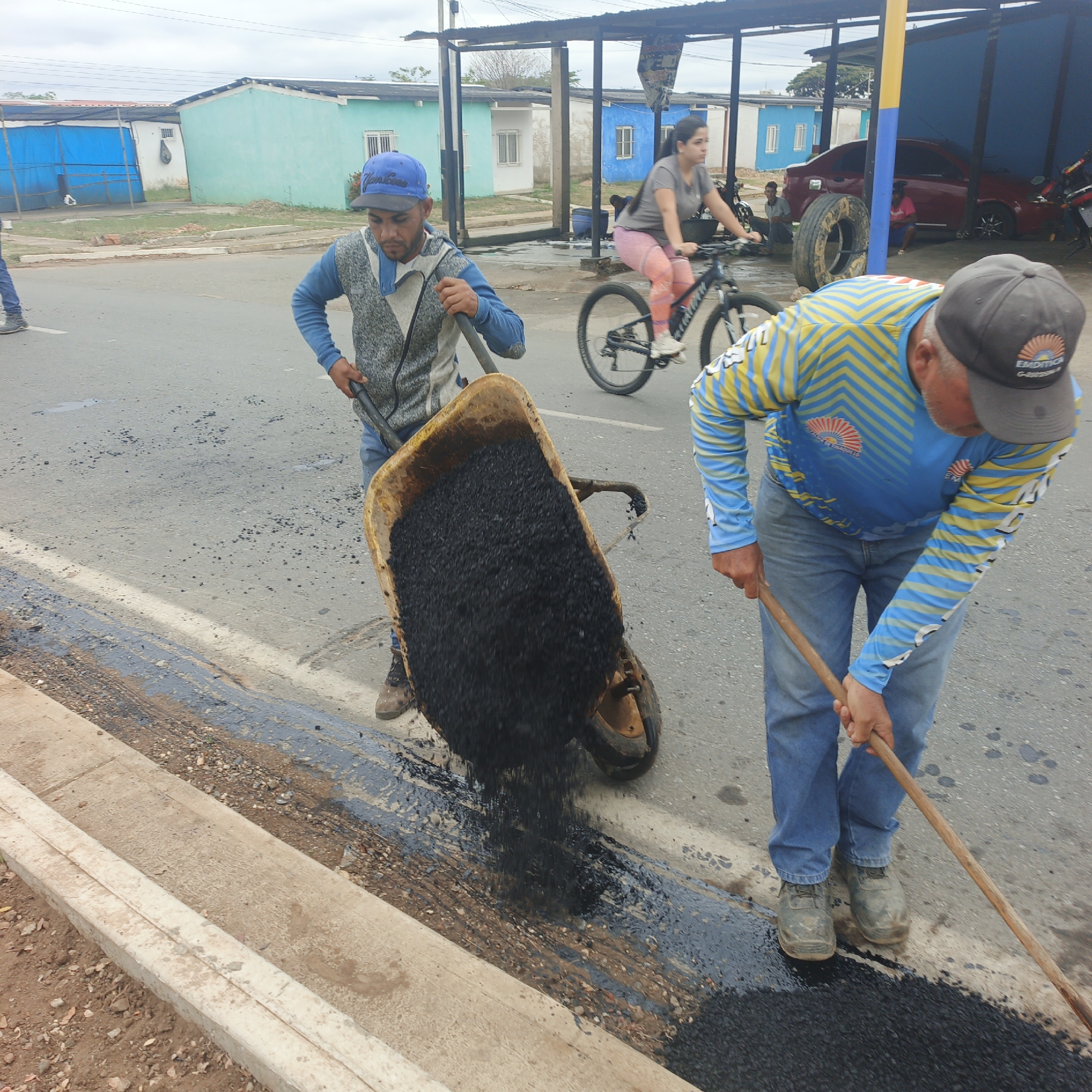 Two men are repairing a street in Tinaquillo, Venezuela. One man is tipping a wheelbarrow filled with asphalt onto the street, while the other is using a wooden tool to spread it. A woman on a bicycle rides by in the background. There are small, brightly painted buildings nearby. A car and several people are under a covered area further in the background.