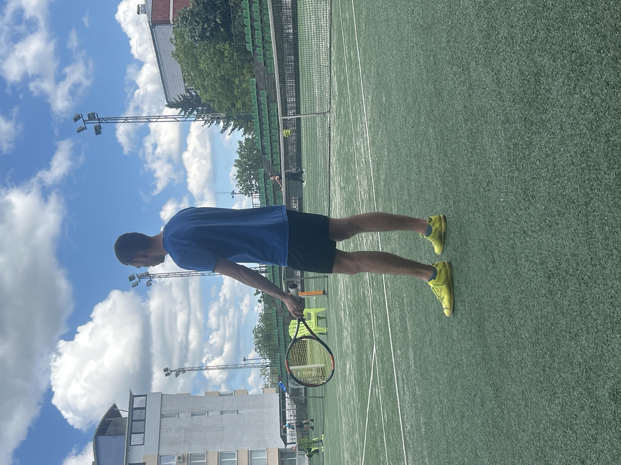 A man in a blue t-shirt, black shorts, and yellow sneakers stands on a tennis court, holding a tennis racket. The court appears to be green hardcourt. A net is visible in the background. Behind the court, there are stands with green seats and trees. Buildings and light posts are visible in the background. The sky is partly cloudy. The location is likely in Veliko Tarnovo, Bulgaria, based on visual characteristics of architecture and foliage typical of the region, though this cannot be confirmed without further information.