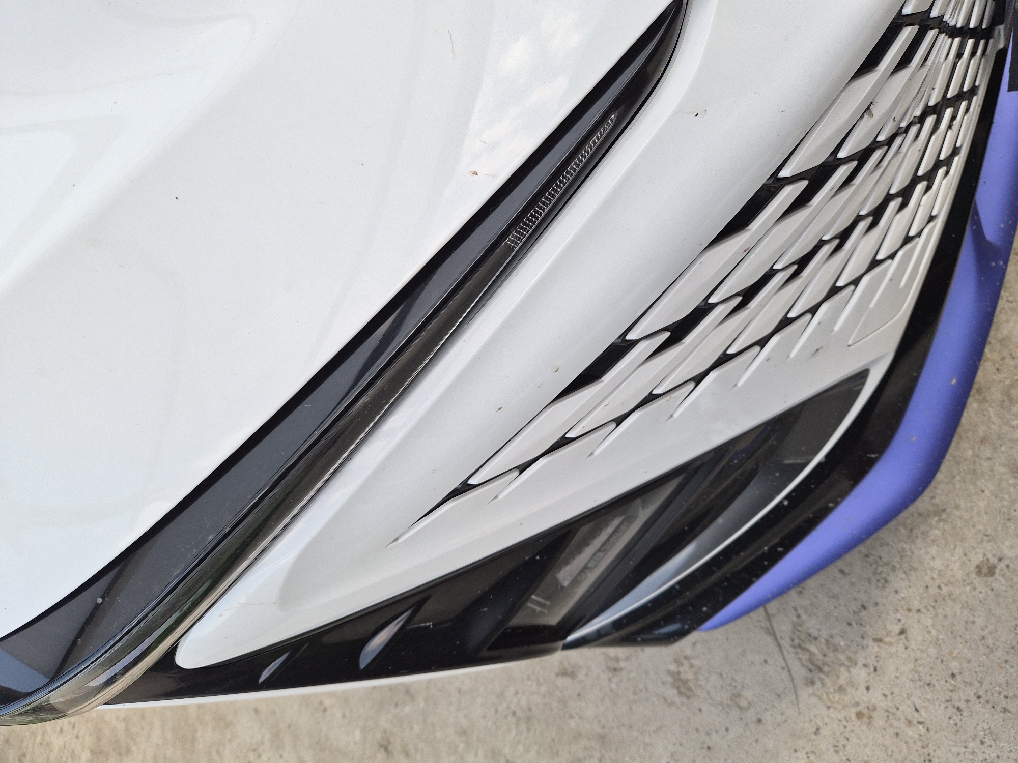 This is a close-up, overhead shot of the front of a white car with sharp, angular design elements. The car's body is predominantly white, with black accents forming a complex, layered design around the grille area. A thin, dark strip with a textured pattern runs diagonally across the upper portion of the grille. Below this, a wider, intricate black and white grille pattern dominates the center, resembling sharp, interlocking teeth or scales. The lower edge of the car features a bright purple trim. The car is parked on a concrete surface, which is visible at the bottom of the frame. There are small specks of dirt and debris on the car's surface, suggesting it is outdoors. The lighting appears to be natural daylight, casting soft shadows. No people are visible. The image focuses solely on the car's aggressive and modern aesthetic. The scene is likely outdoors in a built environment. No visible text is present in the image.