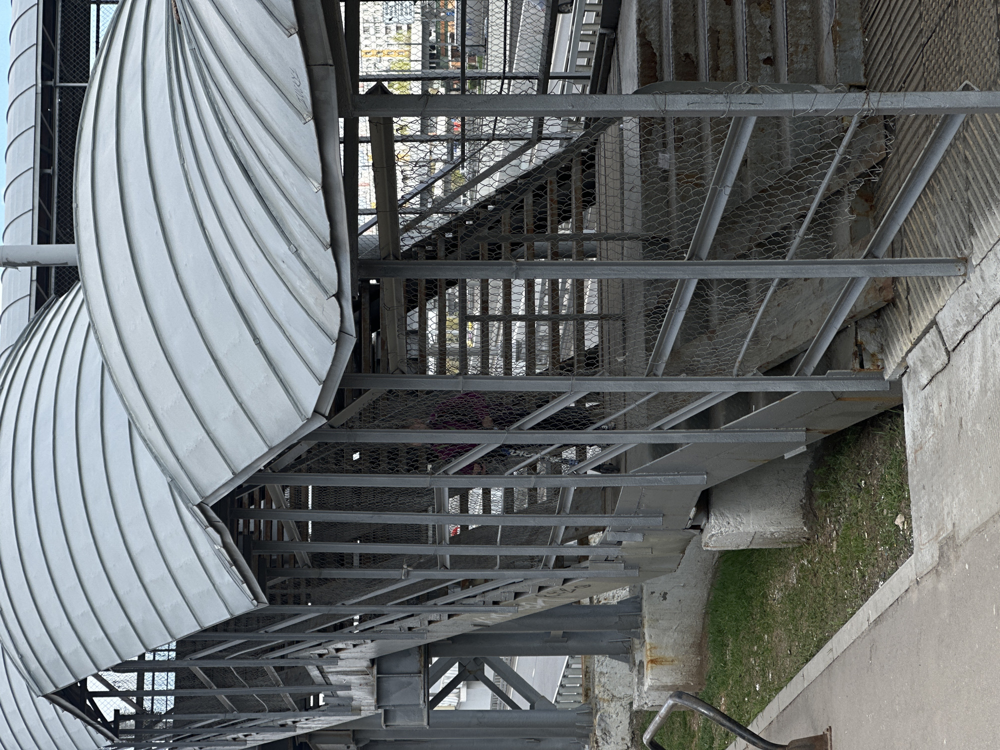 A low-angle shot depicts an exterior metal staircase with a curved, corrugated metal roof. The stairs are caged in wire mesh and supported by a metal frame. Grass and a concrete walkway are visible to the right. A person wearing a purple shirt is on the stairs. Buildings are visible in the background through the wire mesh. The structure is located in Moscow, Russia.