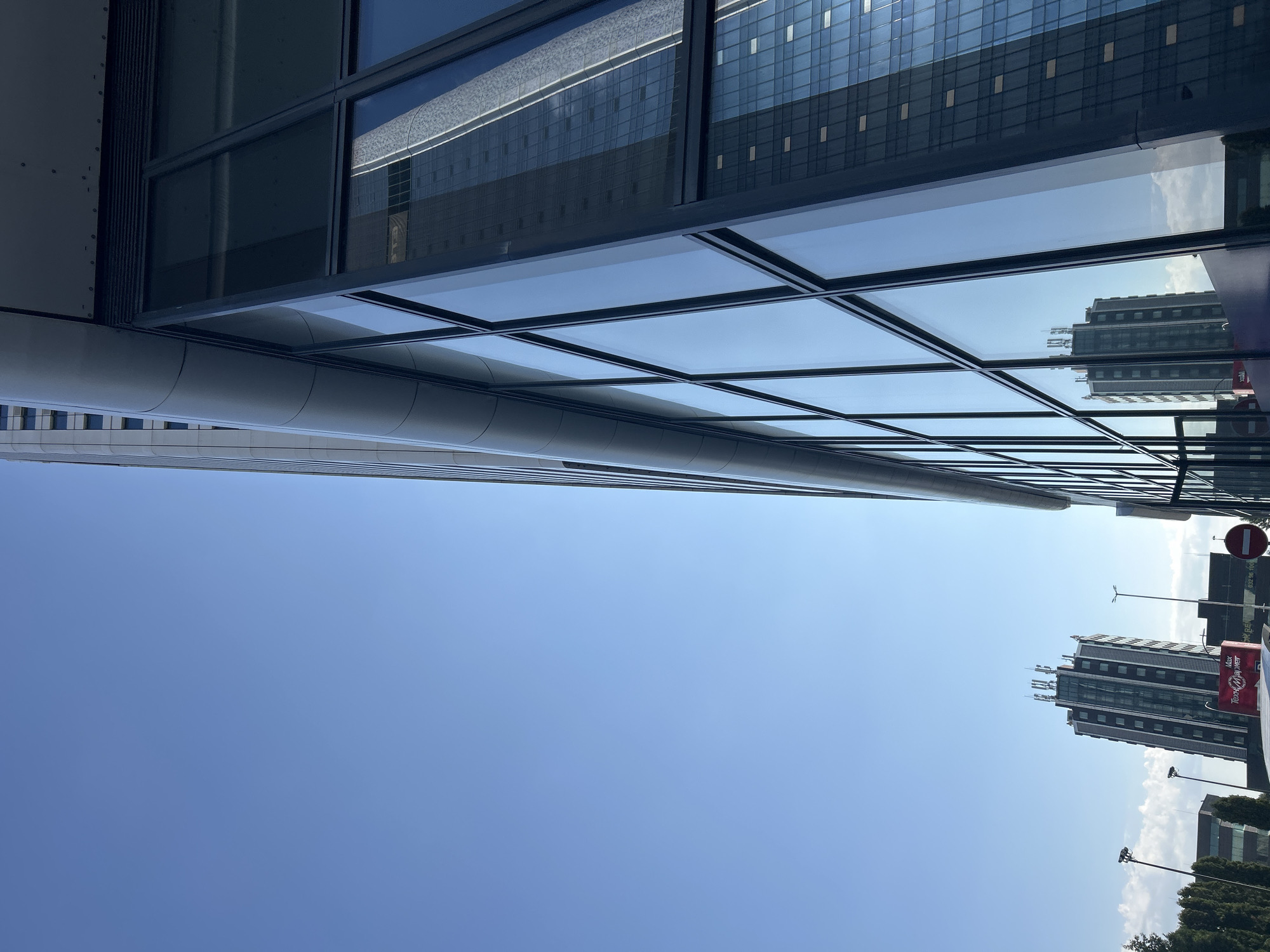 This image depicts the modern architectural style of Sofia, Bulgaria, specifically showcasing the Sky City Business Center and its surroundings. The foreground is dominated by the sleek, glass-paneled facade of the Sky City Business Center, reflecting the clear blue sky and adjacent structures. To the left, the Sky City Business Center itself is visible, a tall, contemporary building with numerous windows and antenna arrays on its roof. Further in the background, other commercial buildings and trees are observable under the bright sunlight. A prominent red banner with white Cyrillic text, likely advertising, is affixed to one of the buildings. The overall scene conveys a sense of urban development and modern infrastructure within Sofia.